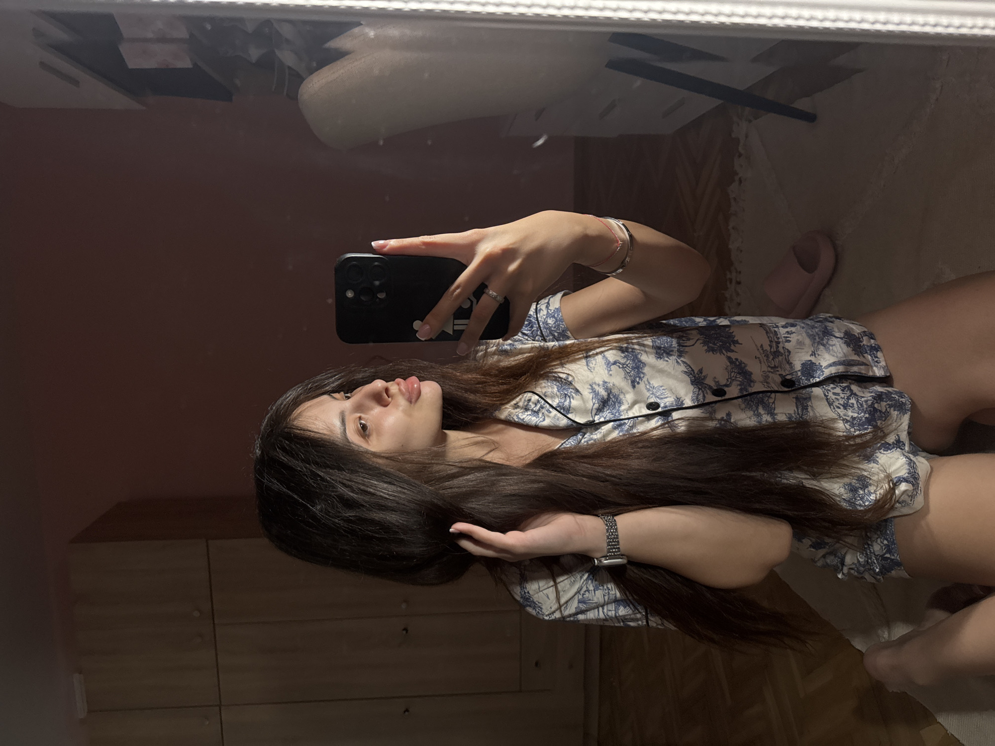 The media file is a selfie taken by a young woman in Plovdiv, Bulgaria. She is lying down, and the camera is positioned to capture her reflection in a mirror. The woman has long, dark hair and is wearing pajamas with a blue and white pattern. She is holding her phone in her right hand, taking the picture. Her left hand is resting on her hair, and she is wearing a watch on her left wrist and a bracelet on her right. Her expression is relaxed and casual.
The setting appears to be a bedroom. The reflection in the mirror shows furniture, including what looks like a dresser or wardrobe with wooden drawers. The room is dimly lit, suggesting it might be evening or nighttime. The flooring is visible in the reflection, appearing to be wooden parquet. A pink slipper is also visible on the floor in the reflection, near the woman's feet. The overall mood is intimate and personal, as is typical of a selfie taken in a private space. There is no visible text in the media file.