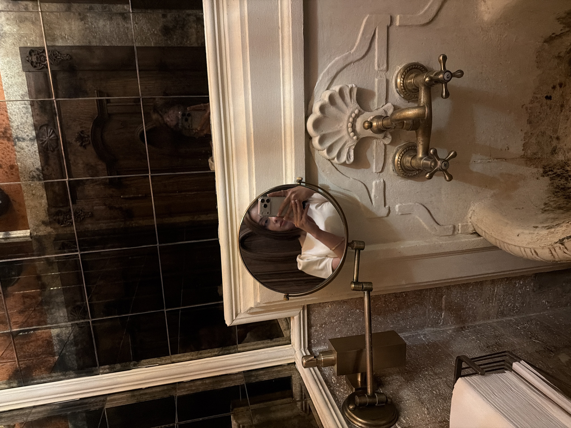 The image shows a bathroom interior. The space features a decorative sink with antique-style faucets mounted on a white, ornate wall. A standing, round mirror is placed near the sink, reflecting a person taking a selfie with a smartphone. The mirror shows their face and part of their white shirt. The reflection in the larger mirror behind shows a grid of dark tiles and a blurry reflection of the scene. A container with stacked white paper towels is present next to the mirror's stand. Yerevan, Armenia is not discernible from the visual information in the image.