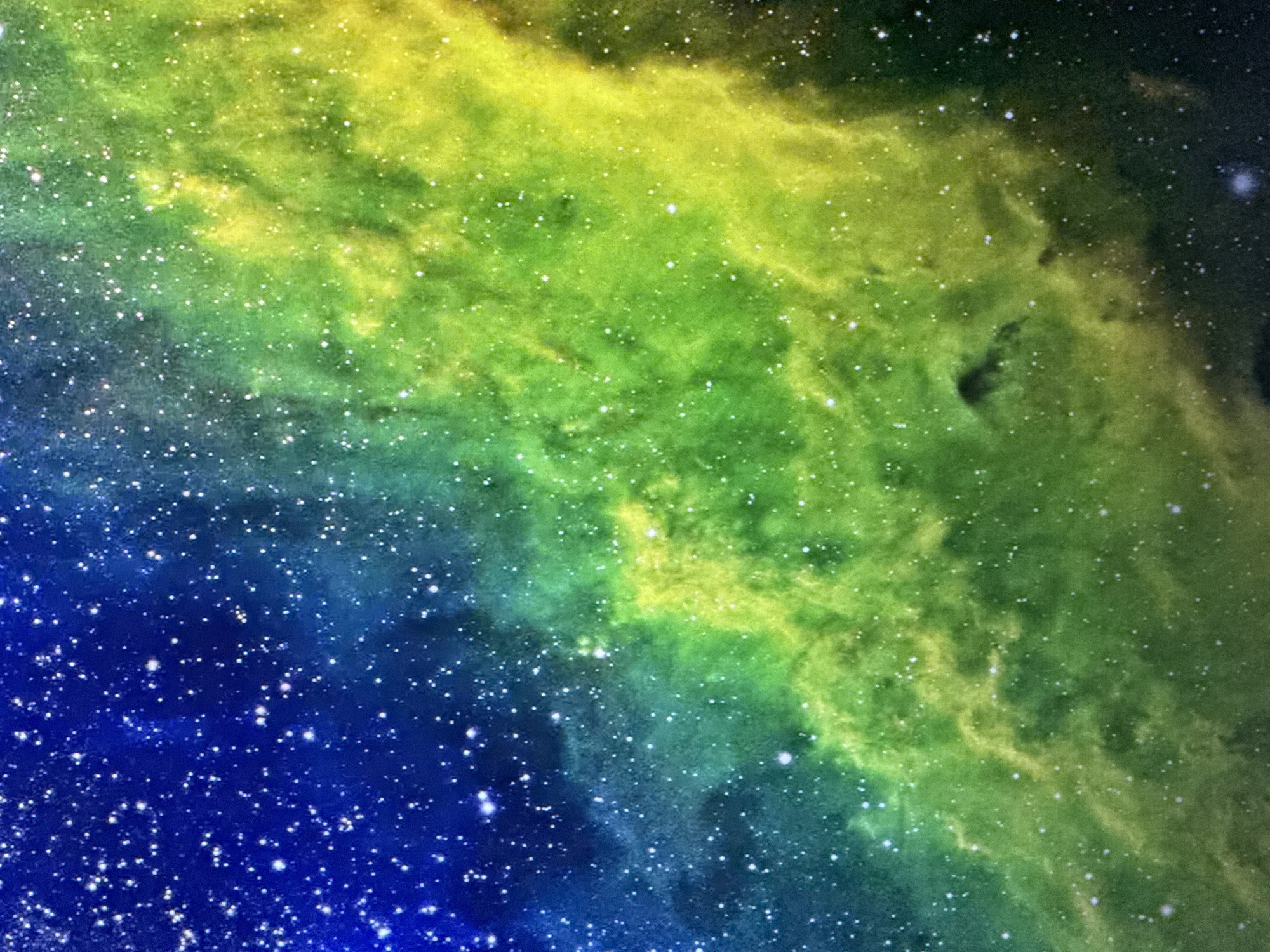 This is a digital image of a nebula in outer space. The nebula is depicted with a gradient of colors, transitioning from deep blue in the lower left to vibrant green and then to yellow in the upper right. Numerous small, white specks are scattered throughout the image, representing stars. The nebula itself has a cloudy, wispy appearance, with variations in density and brightness creating a sense of depth and texture. Some areas are more illuminated, suggesting the presence of brighter gas clouds or intense stellar activity, while other areas are darker and more diffuse. The overall impression is one of vastness and cosmic beauty. There is no text visible in the image.