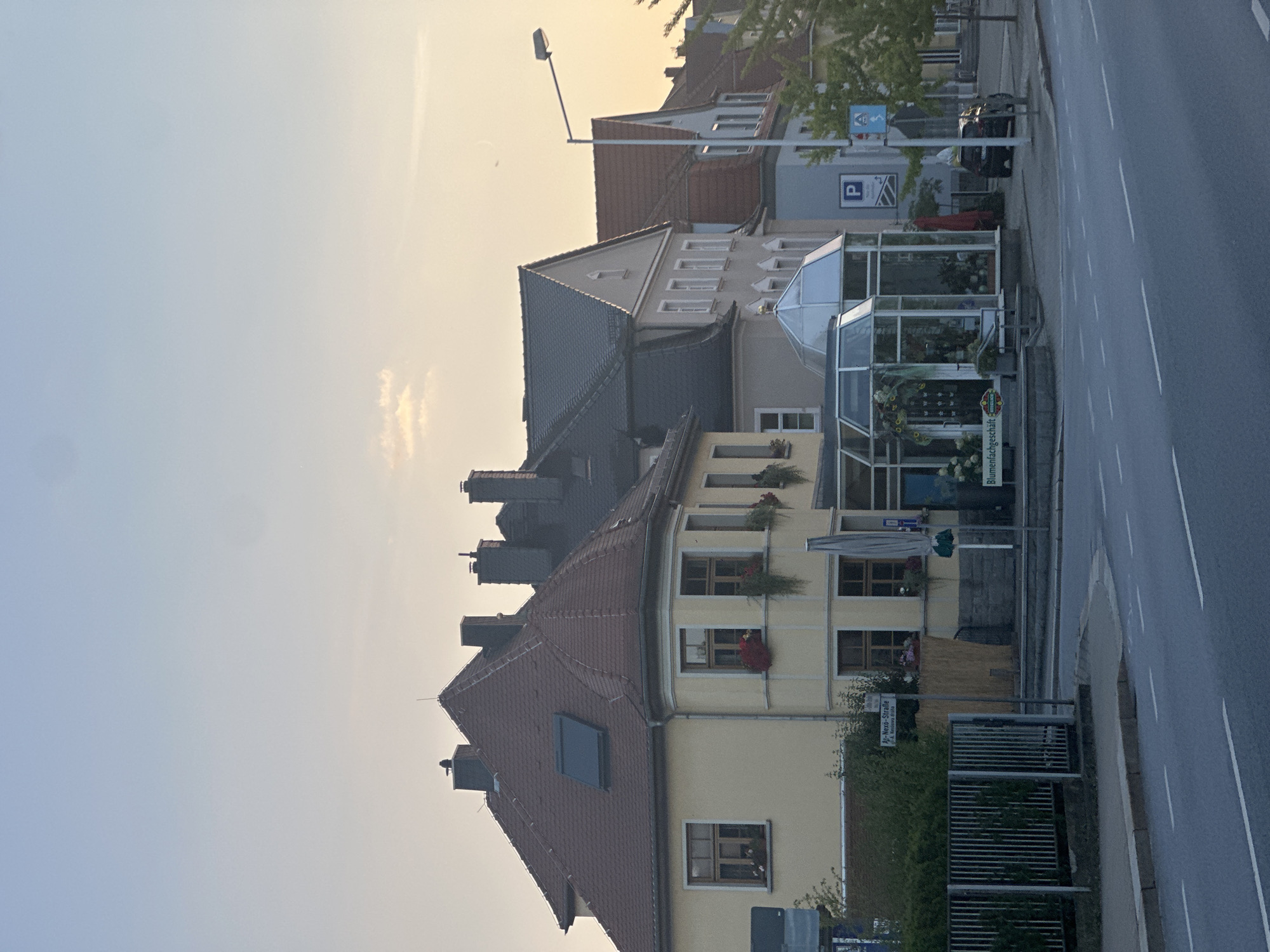 The image shows a street scene in Bautzen, Germany, with several buildings lining the road. The sky is a soft gradient of blue and yellow, suggesting either early morning or late afternoon. On the left, a yellow building with a red tiled roof has several windows, some decorated with flower boxes. A metal fence is visible in front of a green hedge.
Adjacent to the yellow building is a shop with a glass facade, displaying flowers and plants inside. A sign above the entrance reads "Blumenfachgeschäft" which translates to "Flower shop." A dark colored car is parked on the right side of the road, near a parking sign. Further down the street, more buildings are visible, with various roof styles and window arrangements. A street light extends over the road from the right side of the image.
A street sign is visible on a pole with German text, reading "Am Nexe-Straße" and "Dr.-Max-Nixe-Straße." This indicates the location is on Am Nexe-Straße, or Dr.-Max-Nixe-Straße, in Bautzen. The road surface is asphalt with white lane markings. The overall impression is a quiet urban street with commercial establishments.