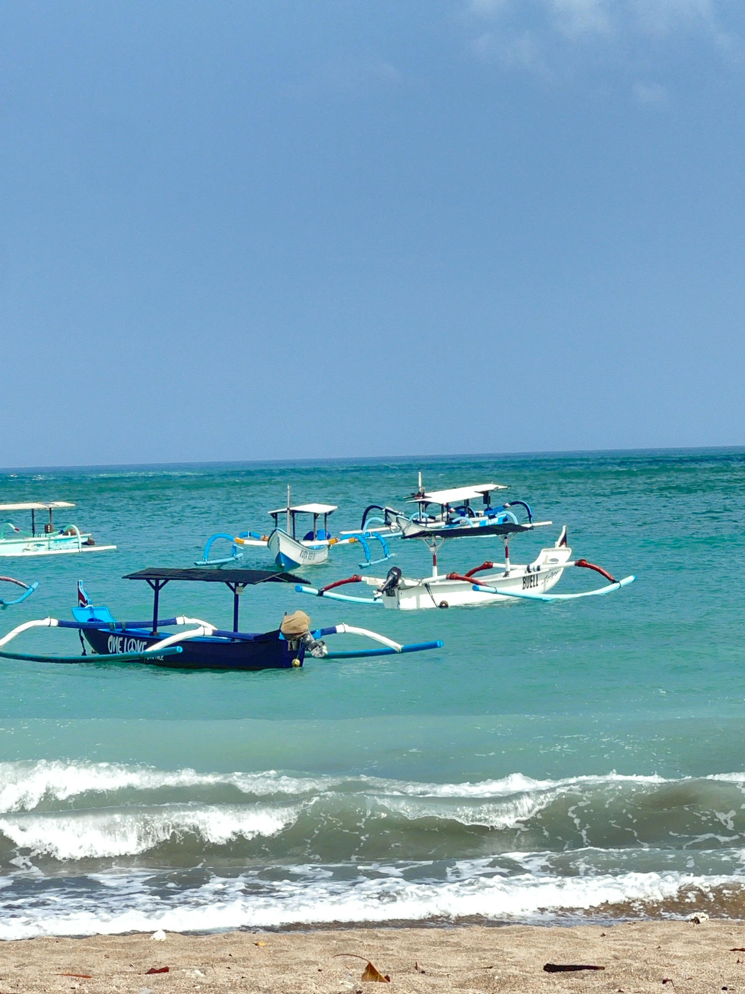 This image depicts a serene scene on the beach in Kuta, Indonesia. Several traditional outrigger boats are anchored in the clear turquoise waters of the ocean. The boats are of various colors, with some predominantly blue and white, featuring the characteristic outriggers that extend from the sides. One boat in the foreground is dark blue with "ONE LOVE" written in white on its side, and has a canopy. Another white boat is closer to the center and has "BUELL" written on it. The sky is a clear, bright blue with a few wispy clouds. The foreground shows a sandy beach with gentle waves rolling in. The overall atmosphere is peaceful and suggests a typical day at a tropical beach destination.