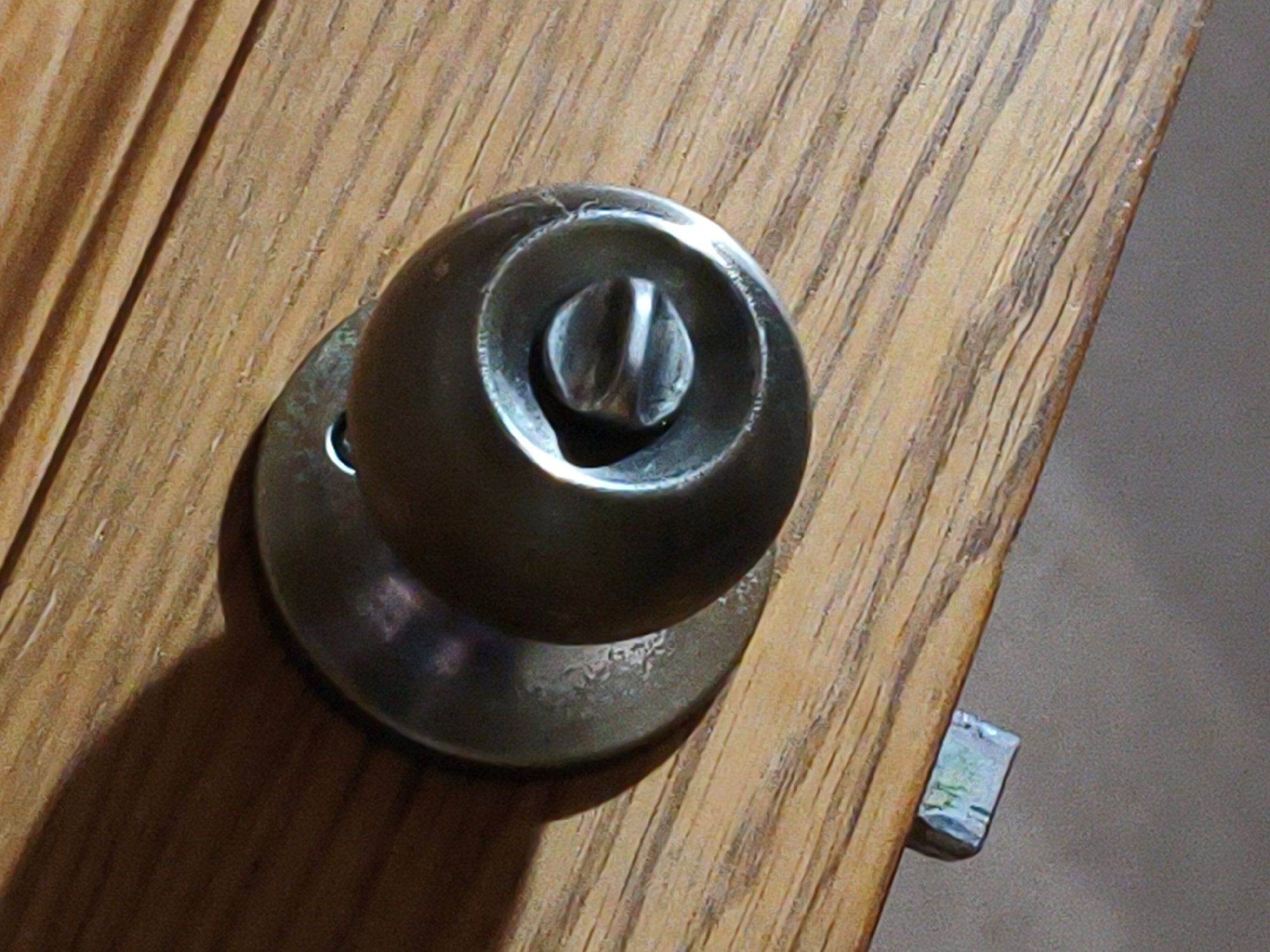 The image shows a close-up, high-angle view of a brushed metal doorknob on a wooden door. The doorknob is round with a slot in the center, and a decorative metal plate is visible beneath it. The wooden door has a visible grain pattern and appears to be made of oak. To the right of the doorknob, a small, rectangular metal latch is partially visible. The lighting suggests an indoor environment. This image does not contain any information that would allow it to be located in Lyubertsy, Russia.