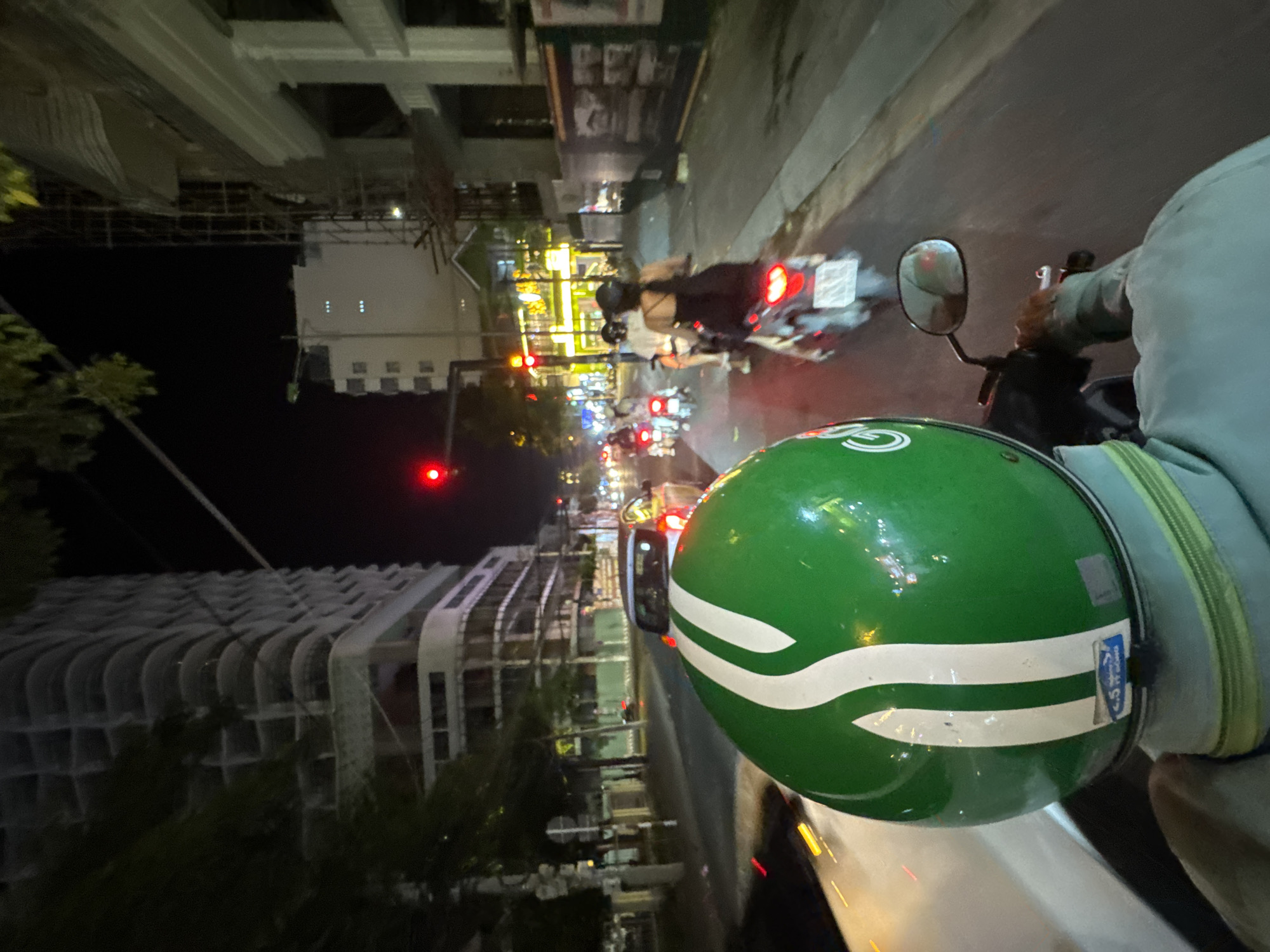 A first-person perspective shot from the back of a motorcycle shows a street scene at night in Nha Trang, Vietnam. The rider is wearing a green helmet with white stripes. In the foreground, the back of the rider's light blue jacket is visible. In the midground, other motorcycles and a car are visible on the road. The road is paved and has painted lane markings. On the right side of the road, there are buildings and streetlights. On the left side of the road, a tall, unfinished building is under construction. The traffic light is red, indicating that the motorcycles and car are stopped. The overall lighting suggests it is nighttime, with artificial lights from buildings and vehicles illuminating the scene.