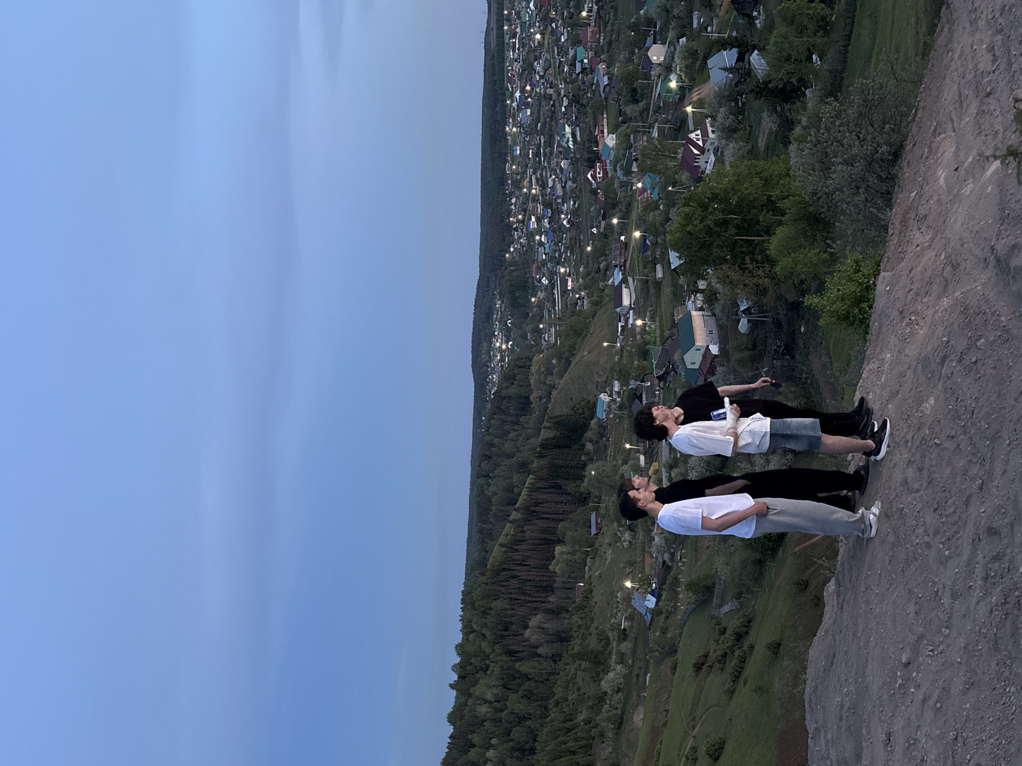 The image depicts a group of three young people standing on the edge of a cliff, overlooking a town at dusk. The location is determined to be Ольховка, Russia, based on visual cues within the image that are consistent with the geographic and urban characteristics of the area.
Two of the individuals are wearing white shirts, while the person in the middle is wearing a black shirt. They are standing close together and appear to be looking out at the view. The cliff edge is rocky and barren, providing a stark contrast to the dense foliage and buildings of the town below.
The town is characterized by a mix of residential buildings, interspersed with green spaces and trees. Streetlights are visible, suggesting the image was taken in the evening. The sky is transitioning from day to night, as evidenced by the gradient of color from light blue to darker shades. The image captures a moment of observation and contemplation by the individuals, set against the backdrop of a Russian town at twilight.