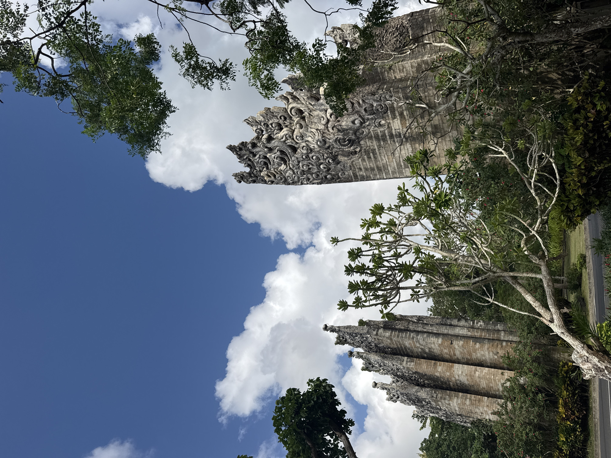 The image shows a view looking up at two ornate stone structures or towers in Kuta Selatan, Indonesia. The sky is visible between the towers, with white clouds and patches of blue. The towers are decorated with intricate carvings. Green trees surround and partially obscure the base of the towers. A road is visible at the bottom right of the image.