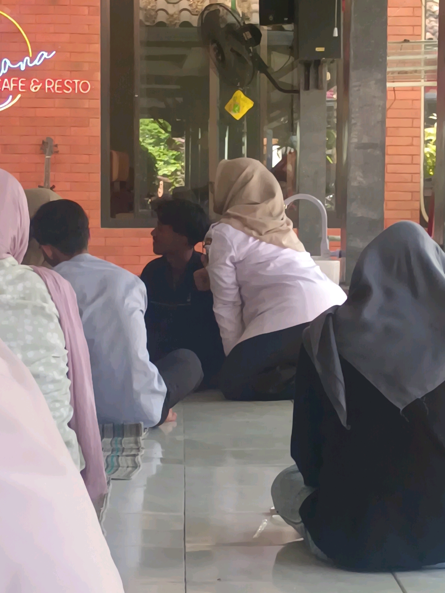 A group of people are sitting on a tiled floor, indoors. In the foreground, a person wearing a black hijab is facing away from the camera, with their back to the viewer. To their left, a person in a light brown hijab is facing the camera and is wearing a light pink shirt and dark pants. To the left of this person, another individual wearing a light blue shirt is also sitting. On the far left, the back of a person wearing a light pink hijab is visible. The background consists of a brick wall with a neon sign that says "ANA CAFE & RESTO" in yellow and pink. A fan is mounted on the wall, and a yellow object is hanging from it. Sunlight streams in from the right side of the image. The scene appears to be in Kerangkulon, Indonesia.