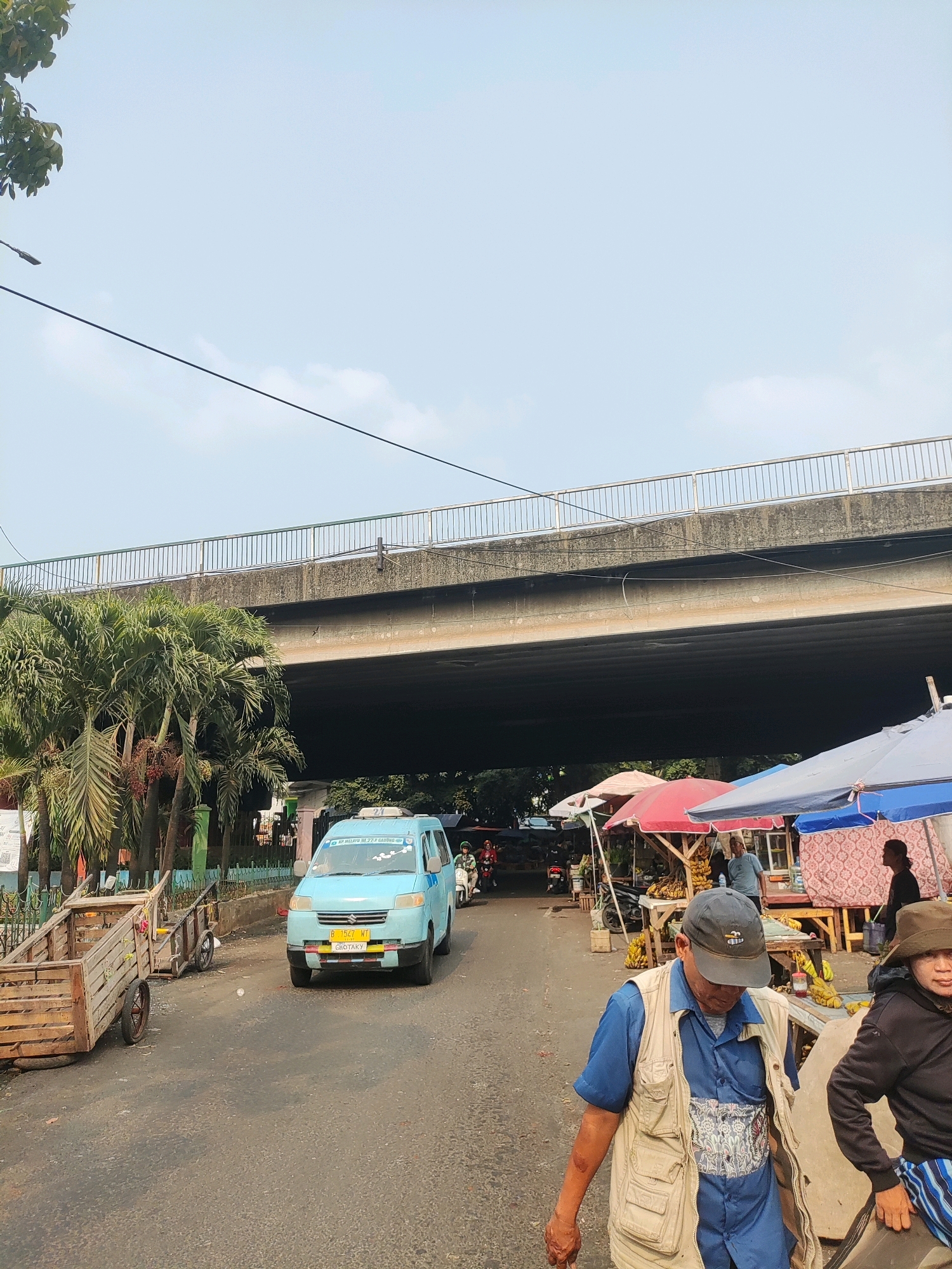 This is a ground-level, eye-level shot taken on a street in East Jakarta, Indonesia, possibly near a flyover or underpass. The sky is a pale blue with some wispy clouds. A large concrete flyover dominates the upper portion of the image. On the left, several palm trees are visible, with some of their fronds reaching up towards the flyover. Adjacent to the trees, a wooden cart with two large wheels is parked. In the center of the road, a light blue van with a license plate "B 1547 WT" is driving away from the camera. The van has some decals on its windshield and a yellow license plate frame. On the right side of the road, there is a market area with several stalls covered by colorful umbrellas (red, blue, and white). Bananas are visible for sale at one of the stalls. Two men are in the foreground, walking towards the right of the frame. The man in the front is wearing a blue shirt, a beige vest with multiple pockets, and a dark baseball cap. He is carrying a light-colored bag across his stomach. The man behind him, to the right, is wearing a dark jacket and a hat. They appear to be walking past the market stalls. The road surface is asphalt and shows signs of wear. There are other vehicles and people in the background, suggesting a busy urban environment.