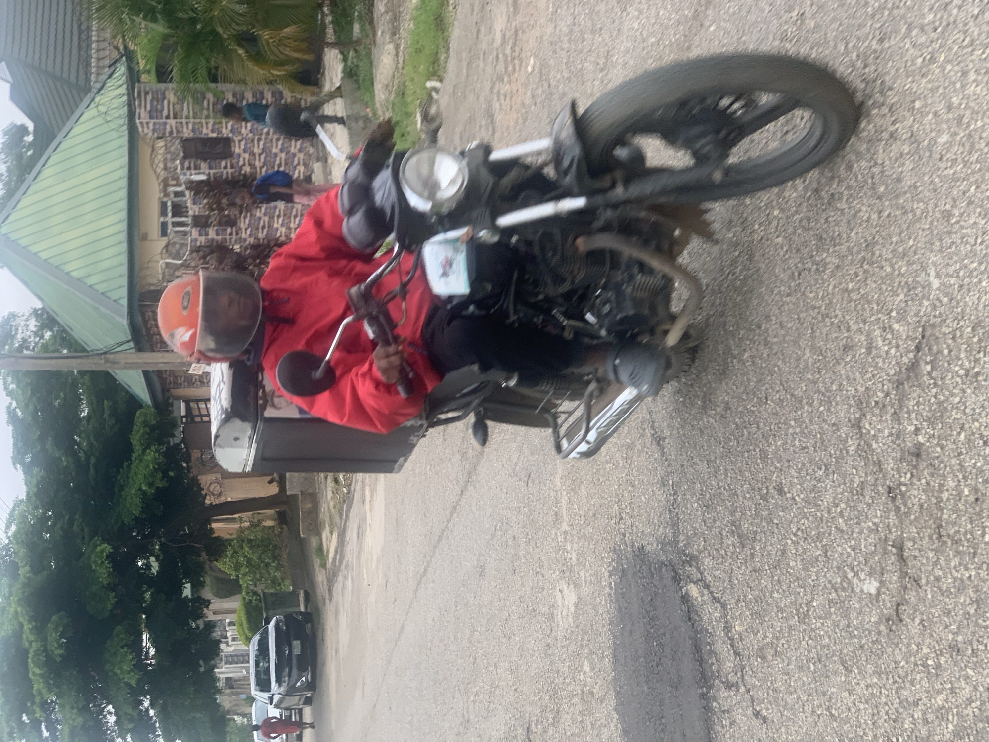 In Abuja, Nigeria, a delivery person wearing a red jacket and an orange helmet rides a black motorcycle with a large black cargo box on the back. The rider is moving forward on a paved road, which appears to be a residential street. The road has some patches of damaged asphalt. In the background, there are houses with green roofs, trees, and other vehicles, including a white car and a black sedan. Two children are visible walking near a house. The scene suggests a typical day in a Nigerian urban environment.