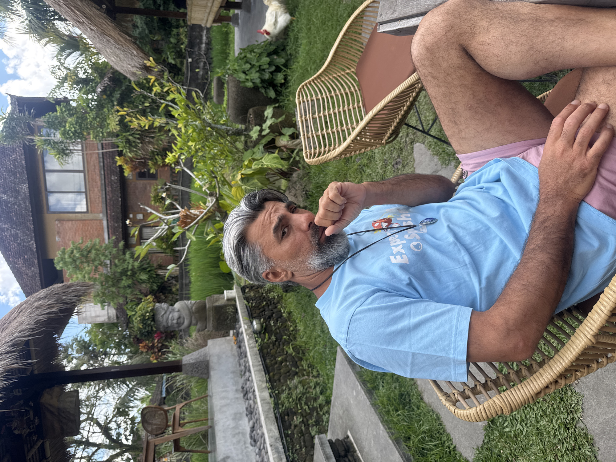 A man with graying hair and beard sits in a rattan chair in an outdoor setting in Tegallalang, Indonesia. He is wearing a light blue t-shirt with text that reads "Experience Bali" and pink shorts. His left hand is raised to his chin, and his right arm is resting on his leg. The background features lush greenery, including rice paddies, a traditional building with a thatched roof, and a chicken foraging on the grass. The scene appears to be a tranquil, tropical environment.