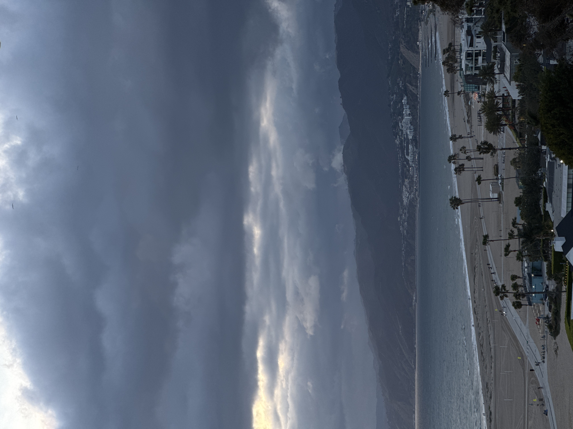 The image shows a panoramic view of Santa Monica, California, USA, under a predominantly cloudy sky. The left side of the image is dominated by heavy, dark storm clouds with streaks of light. The coastline stretches along the right side, where the ocean meets the sandy beach. Rows of palm trees line a path near buildings, including houses and hotels, that are partially visible amidst the trees and landscaping. Mountains can be seen in the background, shrouded in mist. Some people are scattered along the beach and path.