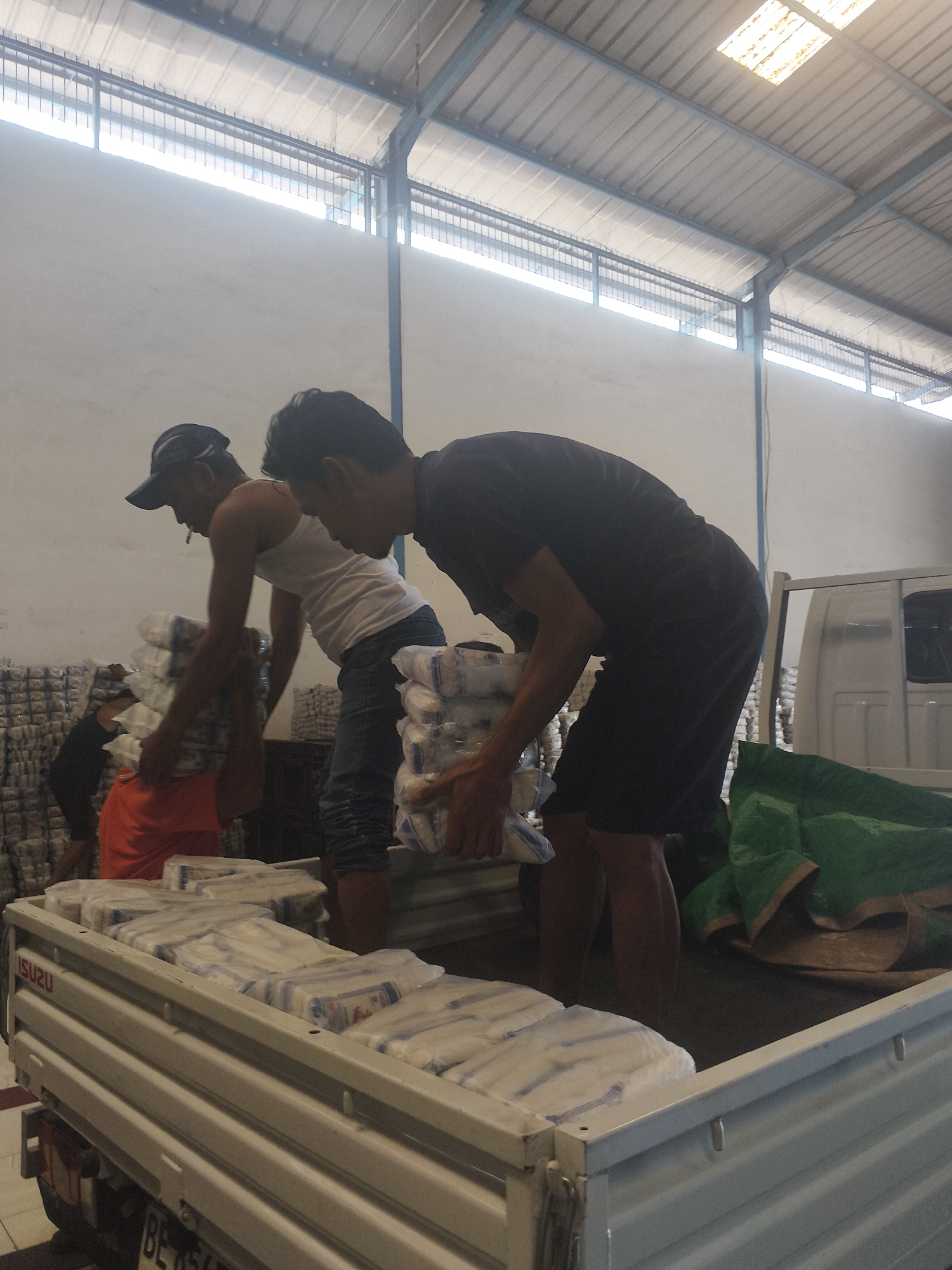 In an industrial warehouse setting in Bandar Lampung, Indonesia, two men are engaged in the physical labor of loading goods onto a white Isuzu truck. The truck's bed is filled with numerous stacks of white, individually wrapped items, possibly detergent or similar packaged products, some covered by a green tarp. The men are actively moving these packages, with one man wearing a white tank top and a baseball cap, and the other in a black t-shirt and dark shorts. The warehouse structure is visible, with a corrugated metal roof and lighting fixtures. The overall scene depicts a logistics or distribution operation.