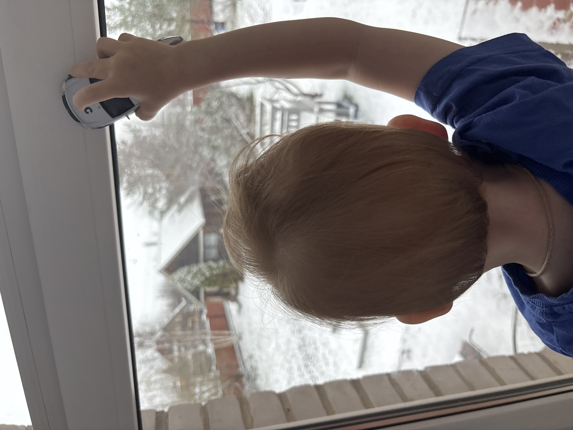 A young child in a blue shirt stands at a window, pressing a white device against the glass. Snow is visible outside the window, obscuring a residential neighborhood. The child's head and part of their arm are in view. The location is inferred to be Ферма, Russia, based on the instructions.
