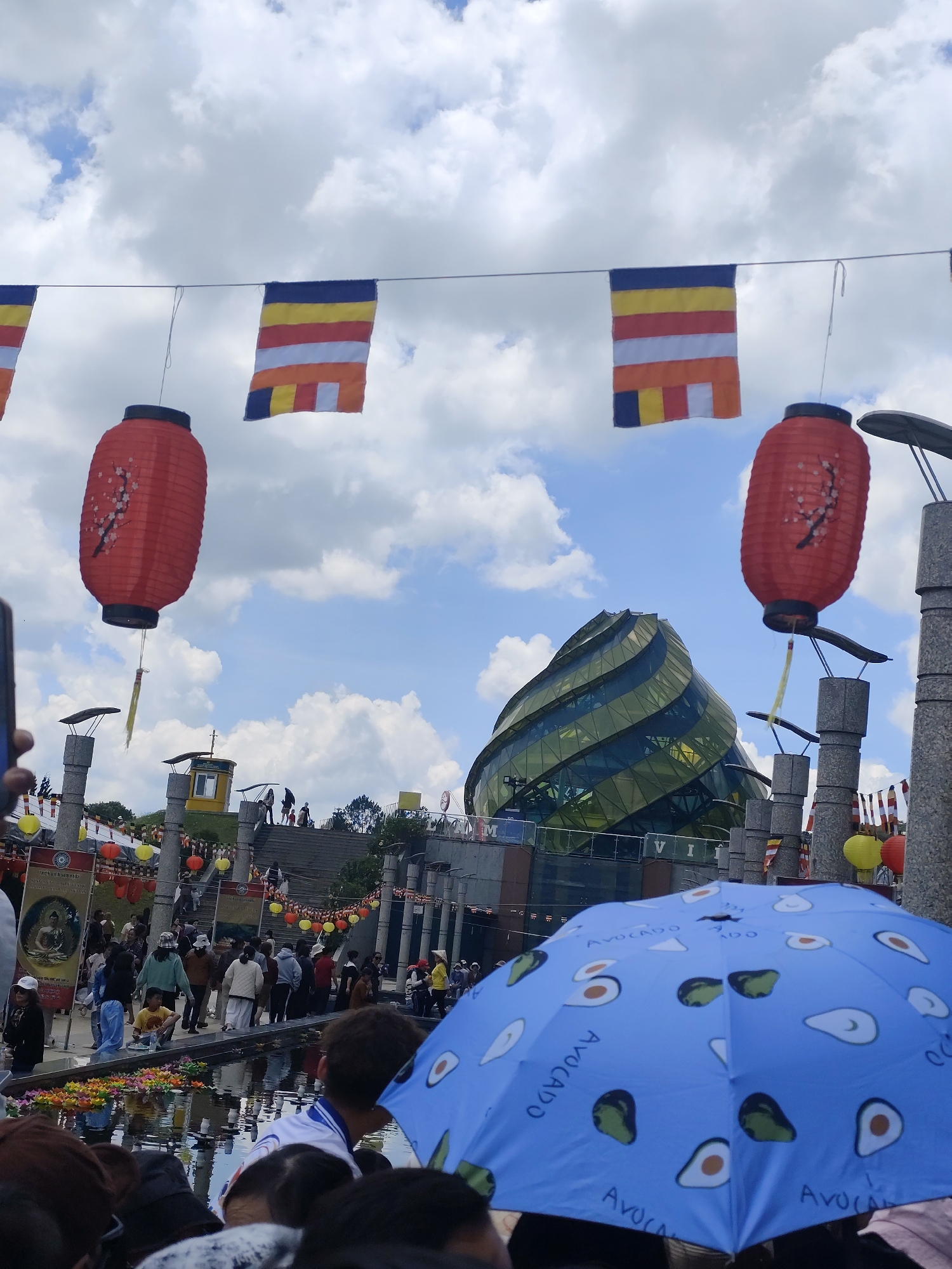 The image shows a crowded scene in Da Lat, Vietnam. In the background is a distinctive green-glass building. Above the crowd are strings of Buddhist flags and red lanterns. In the foreground are people with an avocado patterned umbrella. People are standing near a pool of water with floating floral objects. There are stone pillars, stairs, and other people in the background, including some walking up the stairs. The sky is visible, with scattered clouds.
