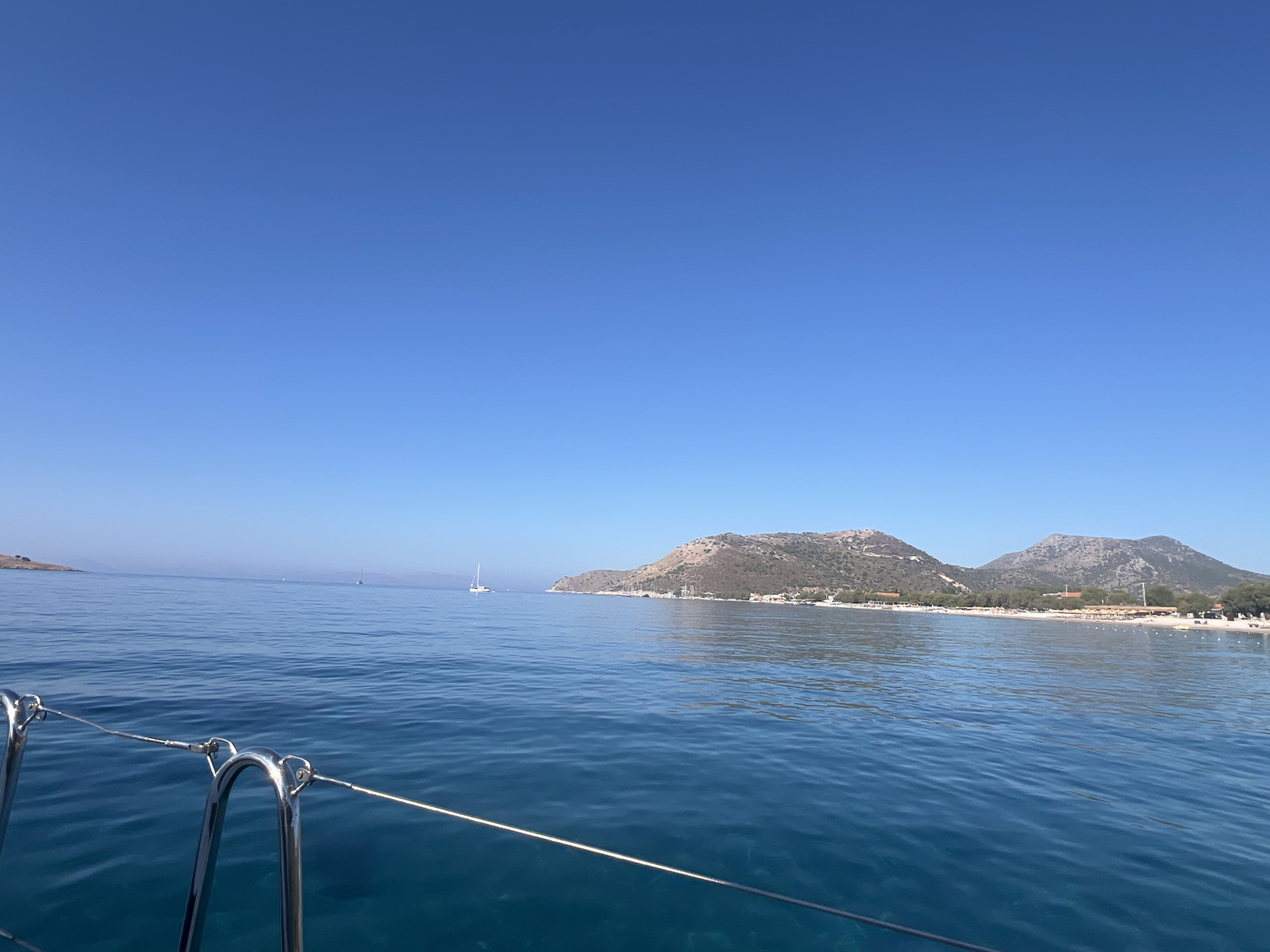 The image captures a serene coastal scene, viewed from the perspective of a moving boat. The foreground features a metallic railing with taut wires, suggesting the boat is equipped with safety features. The sea is a vibrant, deep blue with gentle ripples, indicating calm waters. In the mid-ground, several sailboats are visible, with one white sailboat prominently positioned on the left, and others appearing as distant specks.
The background showcases a picturesque coastline characterized by rolling hills covered in sparse, arid vegetation. A sandy beach stretches along the shoreline, dotted with umbrellas and what appear to be small structures, possibly beachside facilities or houses. The sky is a clear, expansive azure, devoid of clouds, contributing to the overall peaceful and sunny atmosphere. The distant horizon line is clear and sharp, merging the sea and sky. The image composition suggests a leisurely journey along the coast on a bright, clear day.