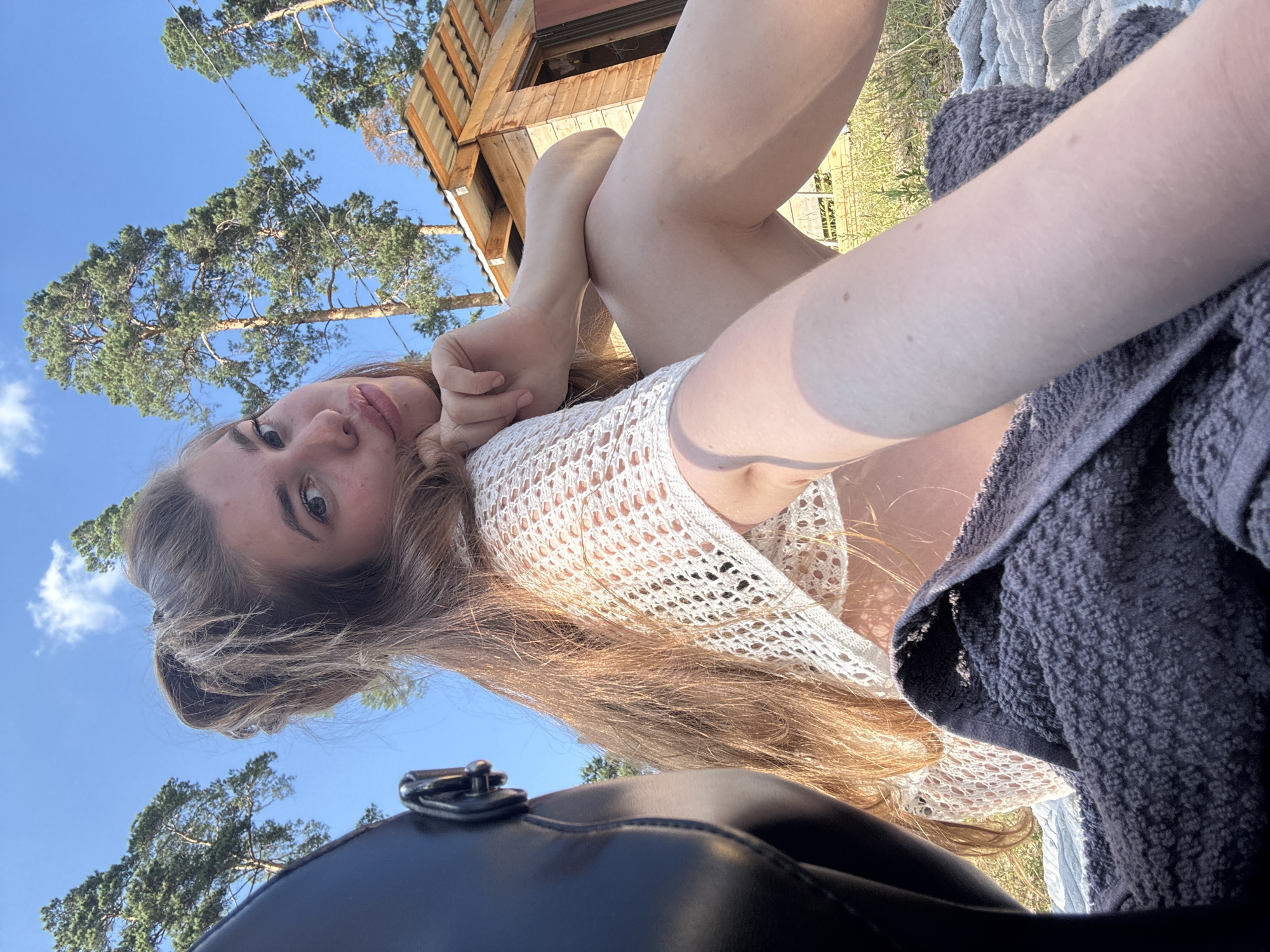 The media file is a low-angle, portrait-oriented selfie taken outdoors. The primary subject is a young woman with long, wavy, light brown hair. She is wearing a white, crocheted top. Her face is partially visible, with her head tilted and her gaze directed towards the camera. She is lying down, with her legs bent and crossed, resting on what appears to be a blanket or towel. The setting is natural, with tall pine trees and a clear blue sky visible in the background. A wooden structure, possibly a treehouse or cabin, is also present in the upper portion of the image. The lighting suggests it is daytime, likely late afternoon given the angle of the sunlight. The overall mood is relaxed and casual. A black object, possibly a bag or backpack, is in the foreground, partially obscuring the view.