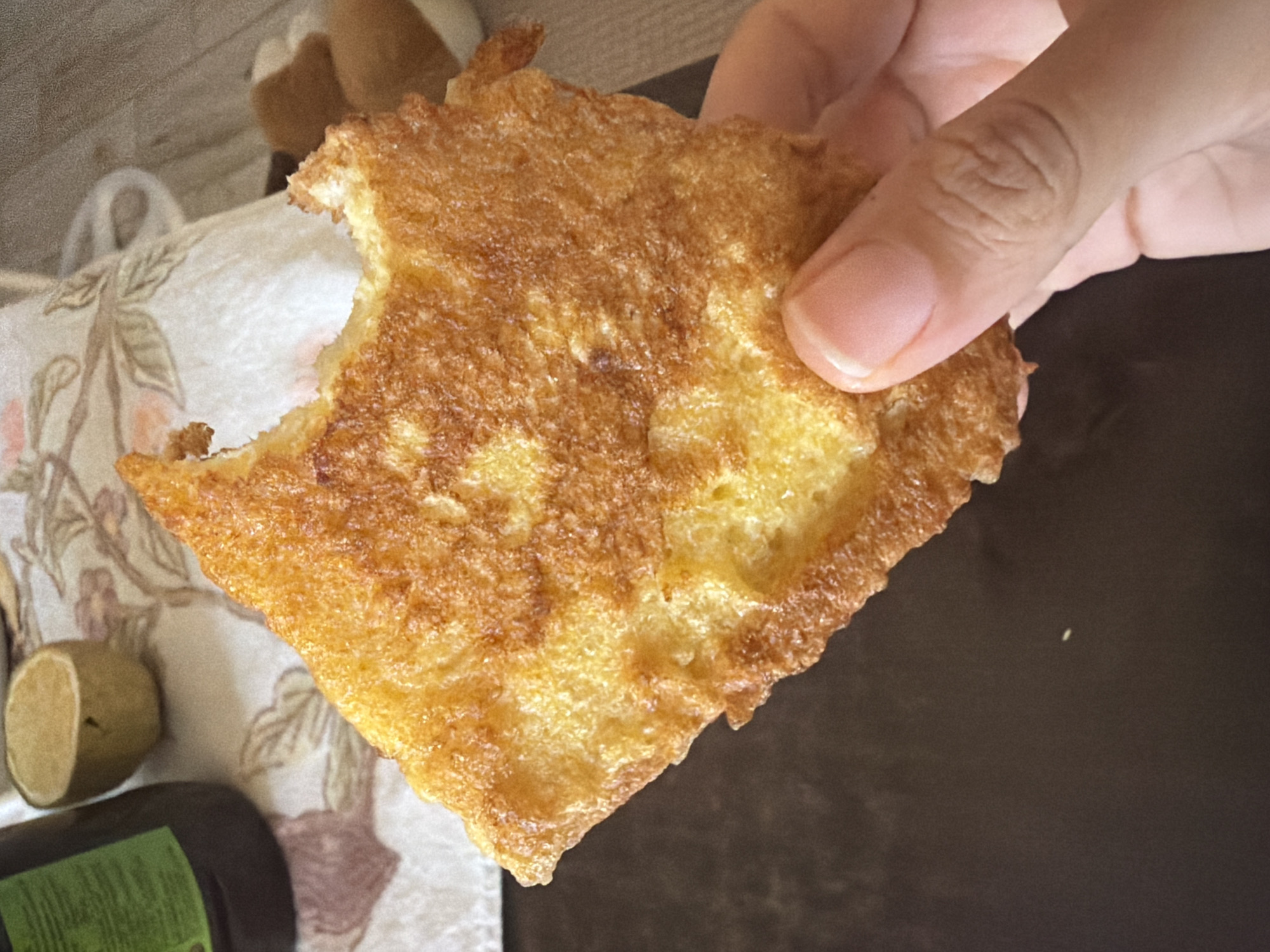 A close-up, slightly angled shot shows a hand holding a golden-brown piece of French toast. The French toast has a bite taken out of it, revealing a fluffy, yellow interior. The toast is slightly greasy, with a crisp, uneven texture. The hand holding the toast has clean, well-manicured fingernails. In the blurred background, a dark bottle with a green label and a halved lemon are visible on a floral-patterned cloth. The setting appears to be a domestic environment, likely in Dromolaxia - Meneou Municipality, Cyprus.