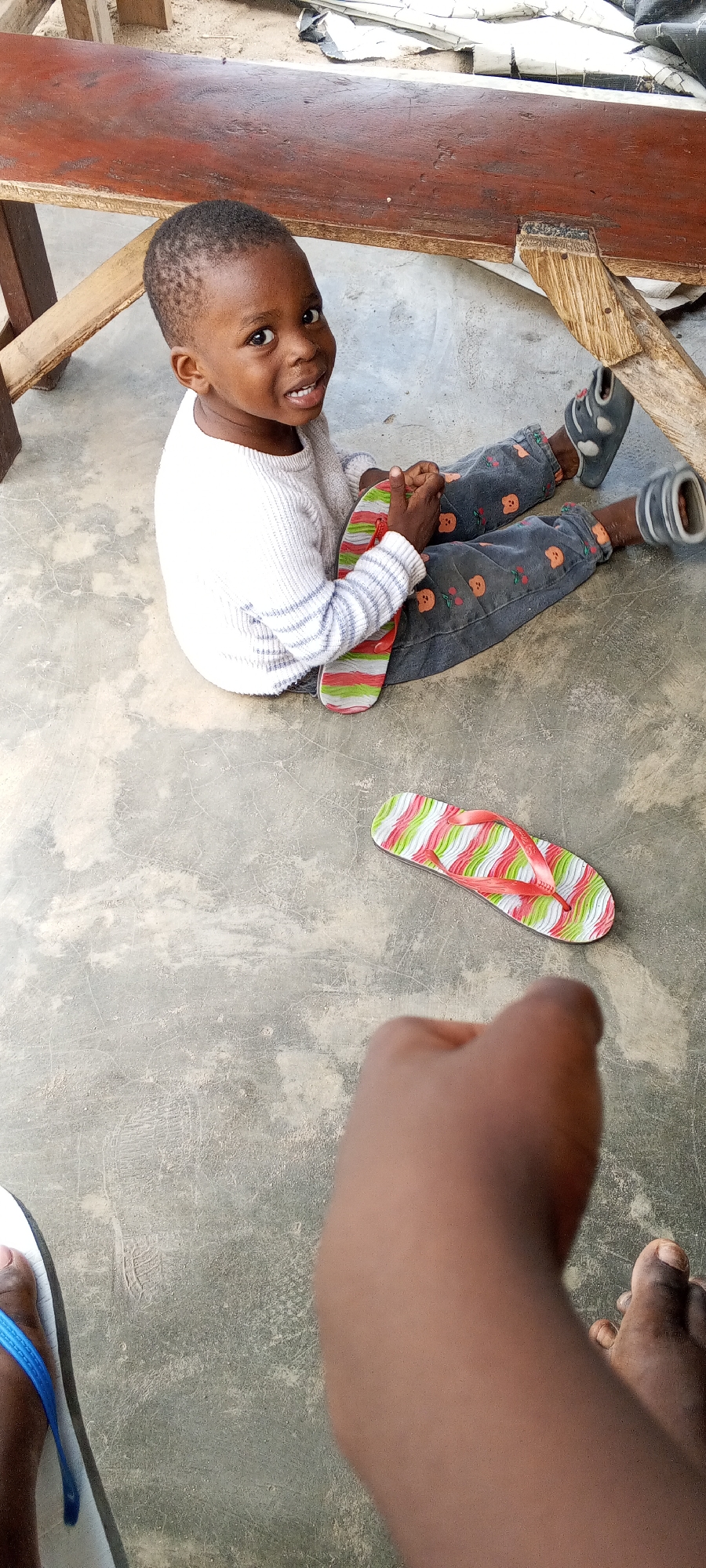 A young, joyful child, likely a boy of African descent, sits on a grey concrete floor in what appears to be a casual outdoor or semi-outdoor setting in Lagos, Nigeria. He has short dark hair and dark eyes, and is smiling broadly at the camera, revealing his teeth. He is wearing a white long-sleeved sweater with light blue stripes on the arms and dark grey pants patterned with small orange cherry or pumpkin-like designs. He wears black closed-toe sandals on his feet.
The child is holding one colorful flip-flop with wavy stripes of red, green, and white. A matching flip-flop rests on the ground nearby. In the foreground, the legs and feet of another person are visible, wearing a white and blue flip-flop, suggesting the perspective of the photographer or someone sitting opposite the child.
Behind the child is a weathered dark brown wooden bench or table with visible wood grain and sturdy wooden legs. Beyond the bench, some light-colored debris or fabric is discernible. The concrete floor shows cracks and dirt marks, indicating its well-used nature. The scene is brightly lit, suggesting daytime. The child's direct gaze and radiant smile convey a cheerful and heartwarming emotion.