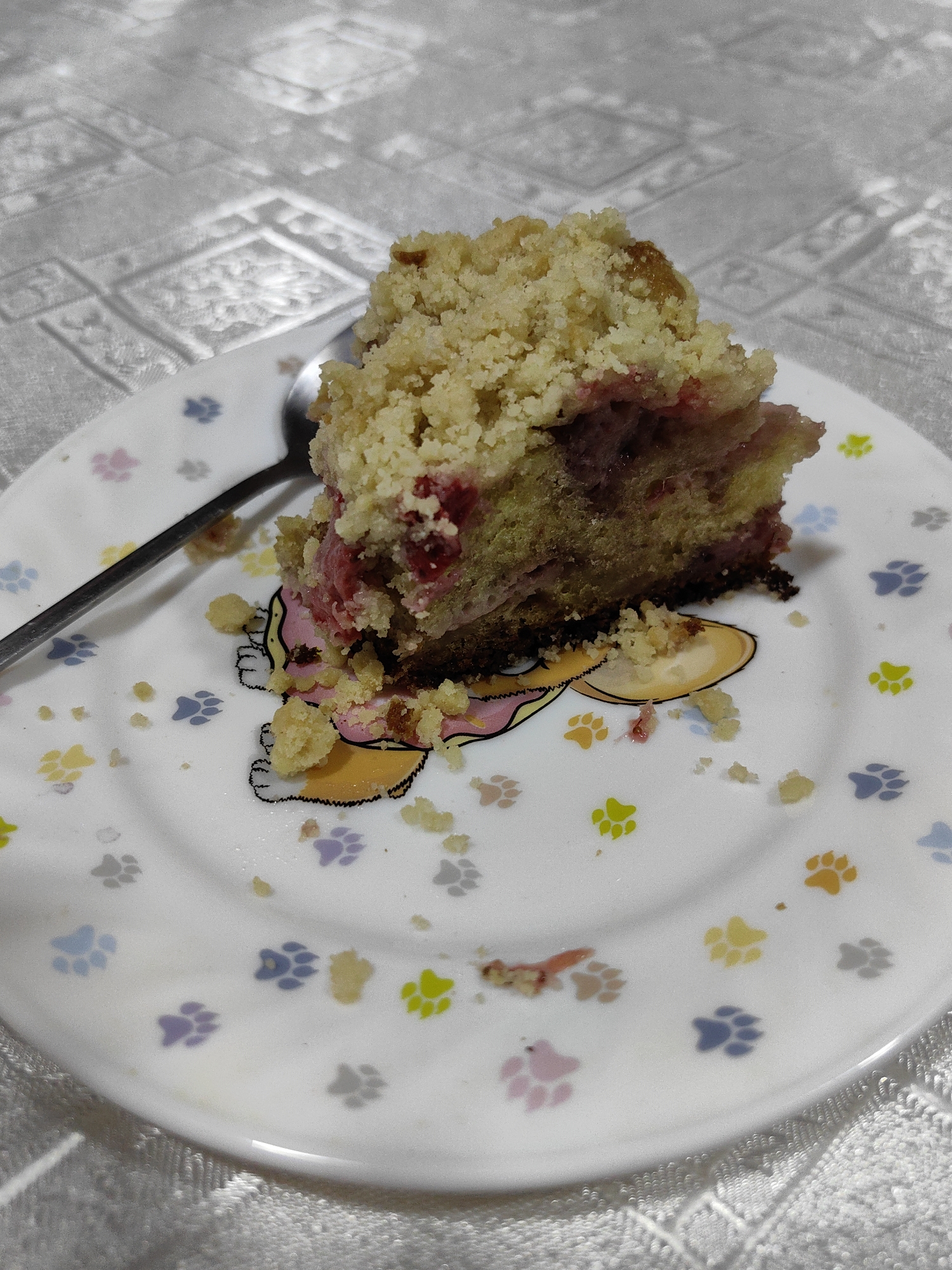 A close-up, overhead shot shows a slice of crumb cake on a white plate decorated with colorful paw prints. The cake, with a crumbly topping and pinkish filling visible, is slightly off-center to the right. A silver spoon rests on the plate to the left of the cake. Scattered crumbs surround the cake slice and are sprinkled across the plate. The plate sits on a white tablecloth with a textured, embossed pattern. The lighting suggests an indoor setting, possibly during the daytime. The overall scene is casual and domestic, with no visible text.