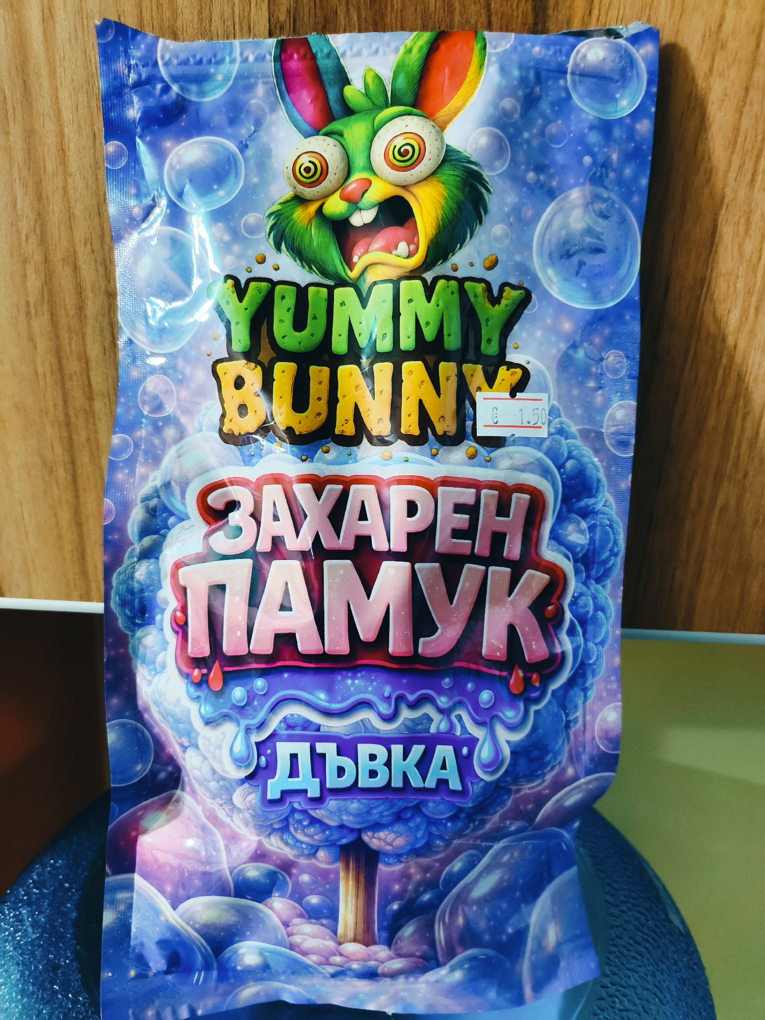 A vibrant, colorful package for a candy or snack product is depicted, resting on a wooden surface. The package features a large, exaggerated cartoon rabbit at the top with bright green fur, pink inner ears, wide, swirly, bloodshot eyes, and an open mouth, conveying a sense of excitement or surprise. Numerous transparent bubbles float across the blue and purple background, which also includes various sparkly elements.
Below the rabbit, prominent English text reads "YUMMY BUNNY" in a textured, bubble-like font, with "YUMMY" in green and "BUNNY" in yellow-orange. A small white price sticker is affixed near the "BUNNY" text, displaying "€ 1.50".
Further down, two lines of Cyrillic text are visible. The upper line, "ЗАХАРЕН ПАМУК," appears in pink and red, outlined in white, and translates to "Cotton Candy." The lower line, "ДЪВКА," is in blue, also outlined in white, and translates to "Chewing Gum" or "Gum." Below this text, the package art shows a whimsical, tree-like structure with a textured, cotton-candy-like canopy in shades of blue and purple, from which blue liquid drips. The base features smooth, bubble-like formations in similar colors.
The setting appears to be an indoor environment, possibly a retail store in Svilengrad, Bulgaria, given the Euro currency on the price tag and the Cyrillic text. The overall design suggests a product aimed at children, with a sweet, fantastical theme.