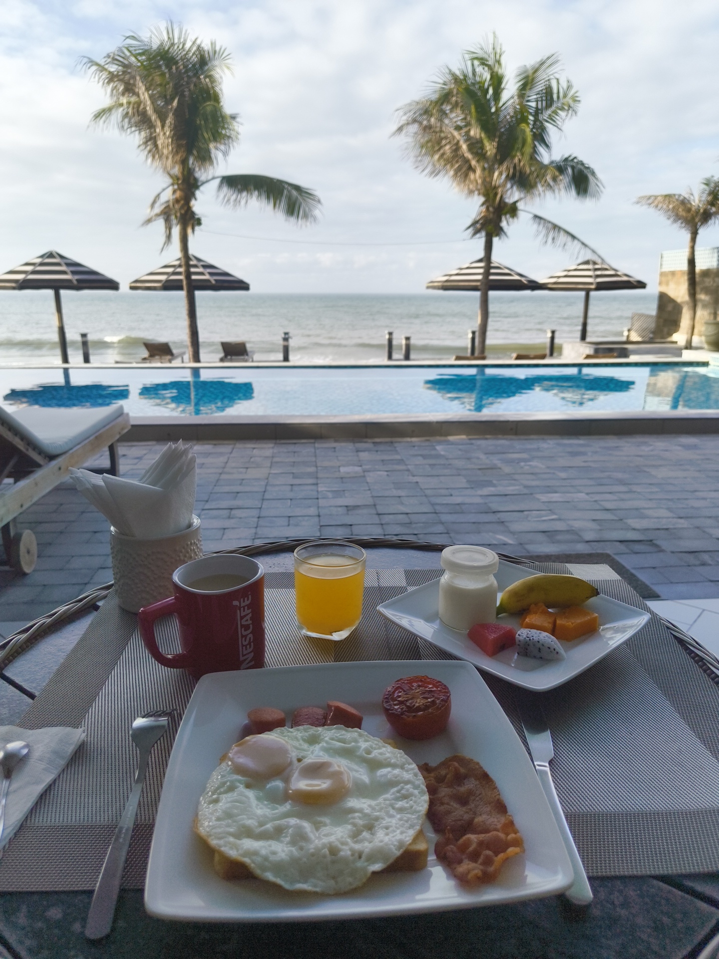 This is a photograph of a breakfast spread on a table overlooking a swimming pool and the ocean in Phu Thuy Ward, Vietnam. The table is set with a placemat, cutlery, a red Nescafé mug filled with coffee, a glass of orange juice, a white plate with a full breakfast including two fried eggs, bacon, sausage, and a grilled tomato. To the right of the main plate, another white plate holds a small glass of yogurt, a banana, and pieces of fruit. In the background, a swimming pool reflects the sky and the surrounding palm trees and umbrellas. Further in the background, lounge chairs are placed on the sand near the ocean. The sky is partly cloudy. There are no people visible in the photograph.