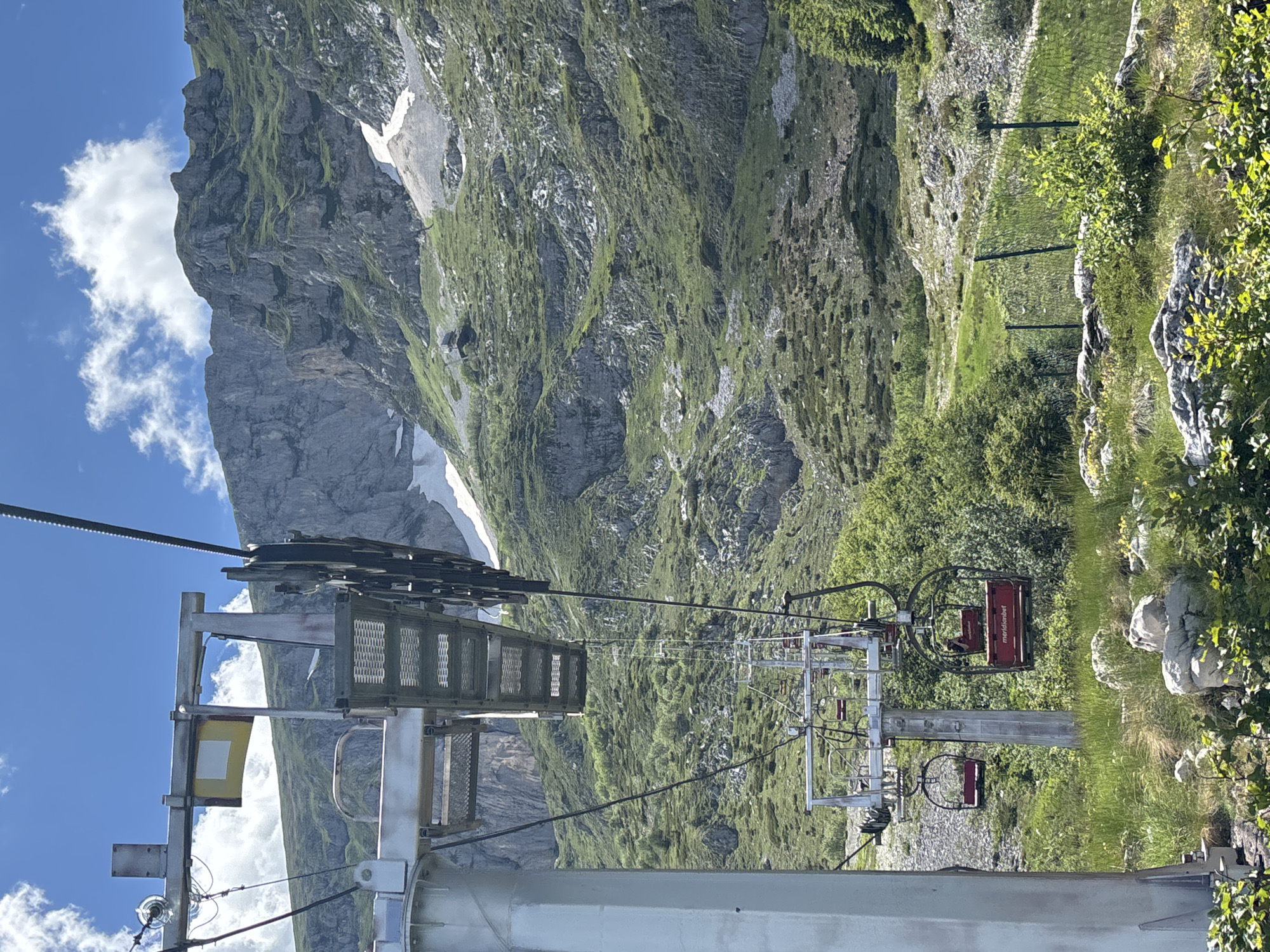 The image depicts a ski lift operating in a mountainous landscape under a clear blue sky with scattered white clouds. The ski lift towers are visible, supporting a cable that suspends red-seated ski lift chairs. The chairs appear to be empty or occupied by unseen individuals. In the background, a steep, rocky mountainside is covered in green vegetation and patches of snow. The terrain is rugged and uneven, with a visible path or track winding through the vegetation. The foreground shows a grassy area with rocks and some low-lying green foliage. The scene appears to be located in Pašina Voda, Montenegro, given the context provided.