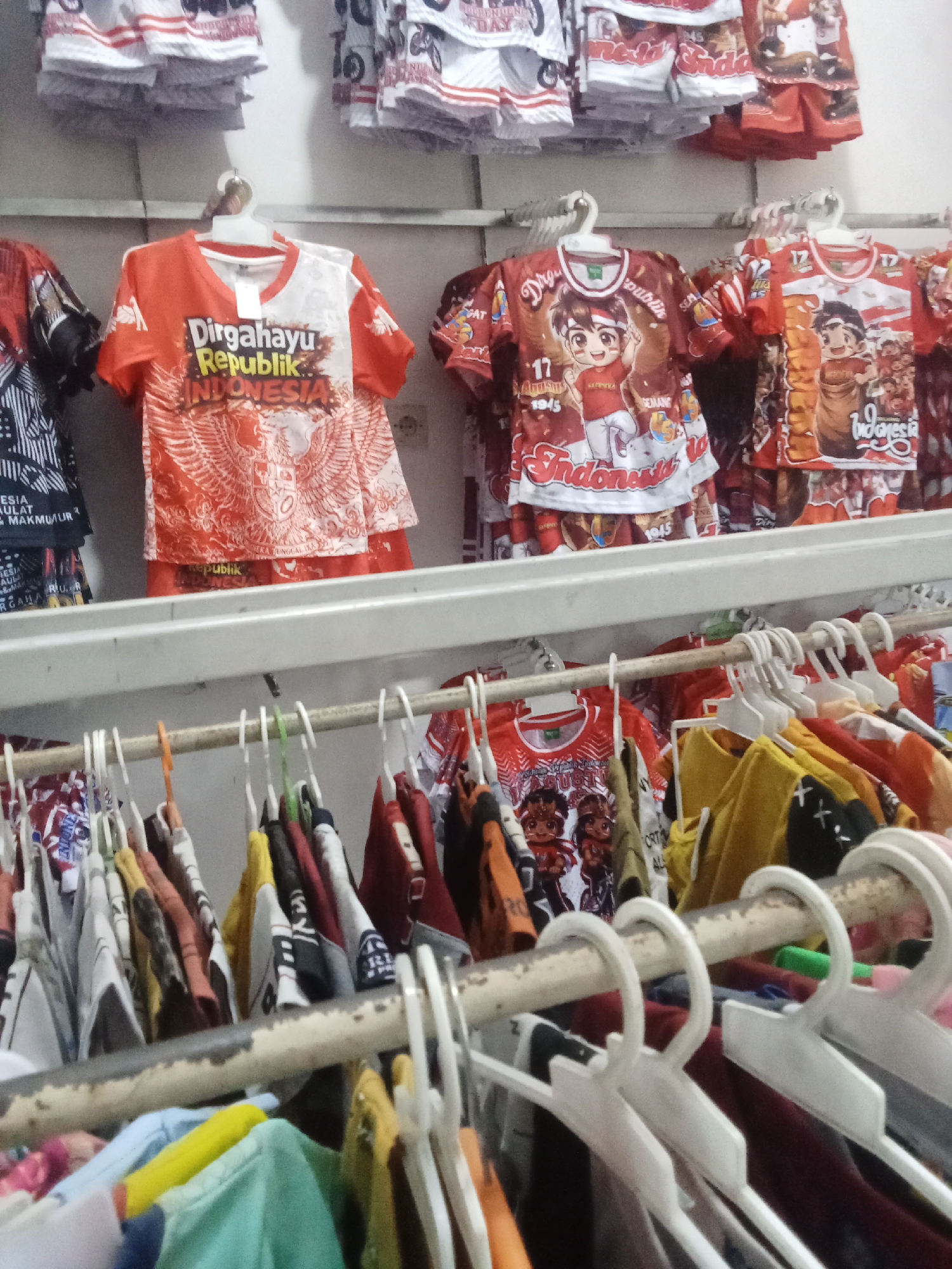 The image depicts a retail setting, likely a clothing store, in Negeri Lama, Indonesia. Rows of t-shirts, predominantly in red and white, are displayed on hangers on a metal rack. Some of the shirts feature Indonesian patriotic motifs, including the text "Dirgahayu Republik Indonesia" and imagery of the Garuda Pancasila emblem, along with cartoon characters. Above this rack, more t-shirts and what appear to be shorts or small garments are stacked. The overall impression is of a merchandise display for sale.