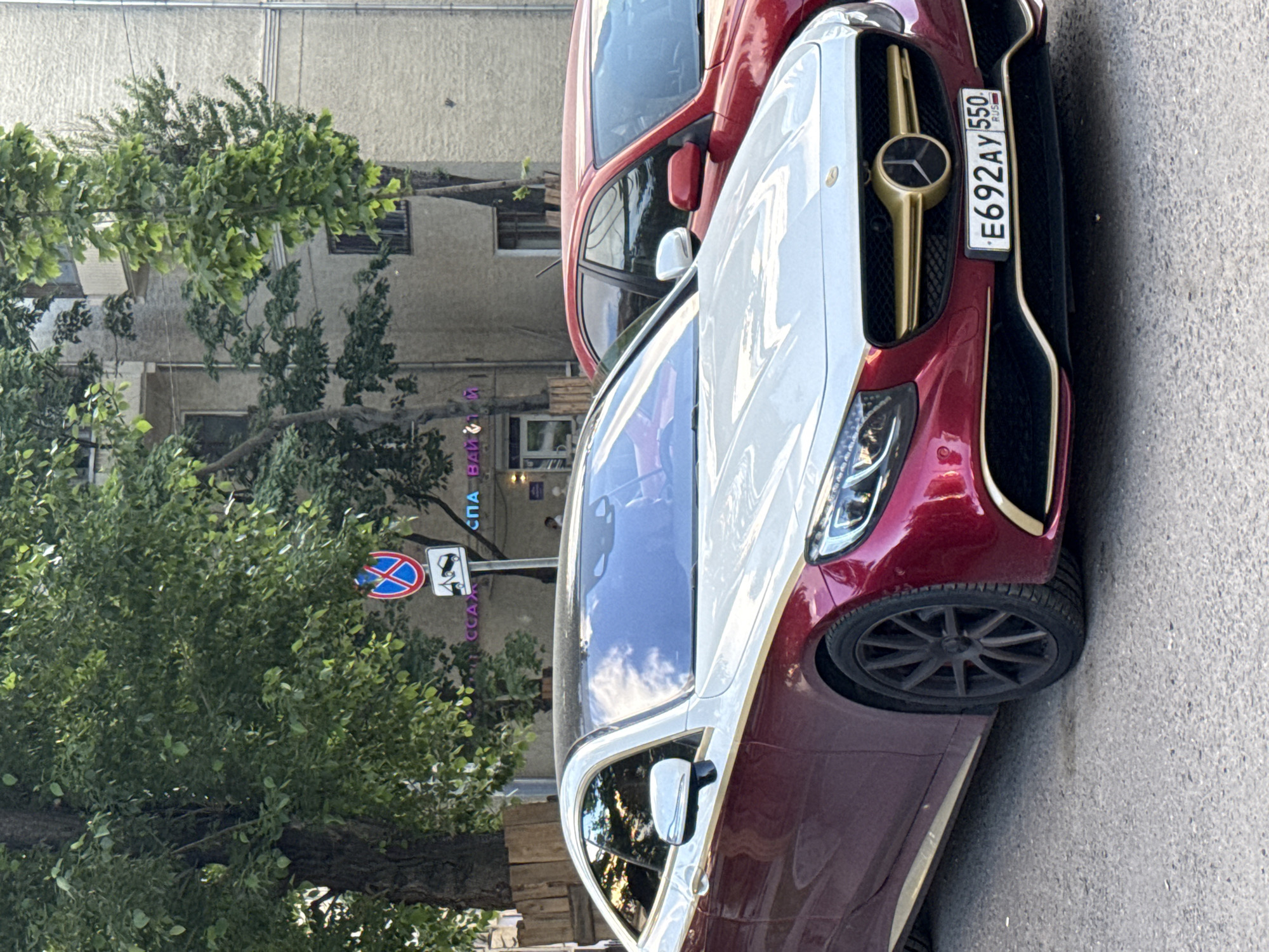 An opulent, two-tone Mercedes-Benz luxury car is prominently featured, parked on an asphalt street in what appears to be an urban setting in Moscow, Russia. The car's lower half is a deep maroon or reddish-purple, transitioning to a light white or cream on the upper body and roof. Its distinctive front grille boasts a gold finish, complete with a matching gold Mercedes-Benz star emblem. Modern LED headlights are visible, and the car is fitted with dark, multi-spoke alloy wheels. A Russian license plate, "E692AY 550 RUS," is clearly displayed on the front.
Behind the Mercedes, a portion of a red car is visible, suggesting a row of parked vehicles. The background consists of green trees with abundant foliage, partially obscuring a light-colored building, possibly an apartment block or commercial structure.
A circular "No Stopping" road sign (blue with a red border and diagonal line) is visible through the trees on the left, beneath which a smaller sign with a tow truck icon indicates enforcement. Faint Cyrillic text, possibly "СПА" (SPA) and "БАНИ" (BANI) with other letters like "CCAX", can be seen on a sign or building in the background.
The scene depicts a daytime setting with clear weather, indicated by the reflections on the car's surface. The cars are stationary, with no specific activity occurring.