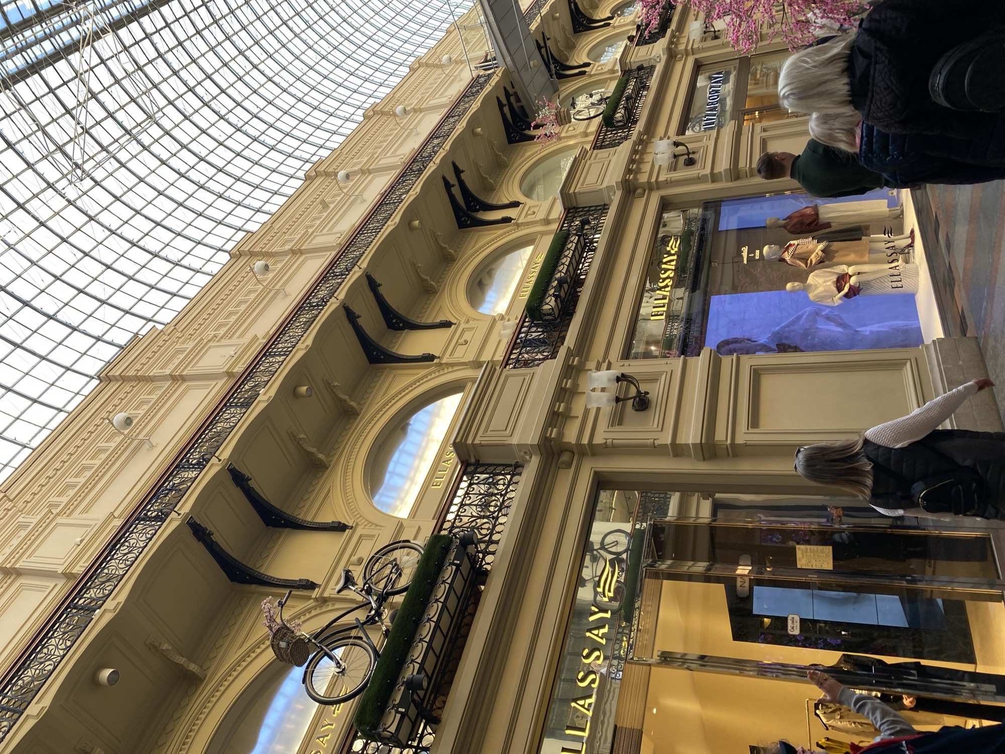 The image depicts the interior of GUM (Main Universal Store) in Moscow, Russia. The architecture includes a glass ceiling and multiple levels with arched windows and decorative details. The store facades are adorned with the name "ELLASSAY." There are bicycles with flower baskets mounted on balconies. People are walking around and looking at displays, and mannequins are visible in shop windows.