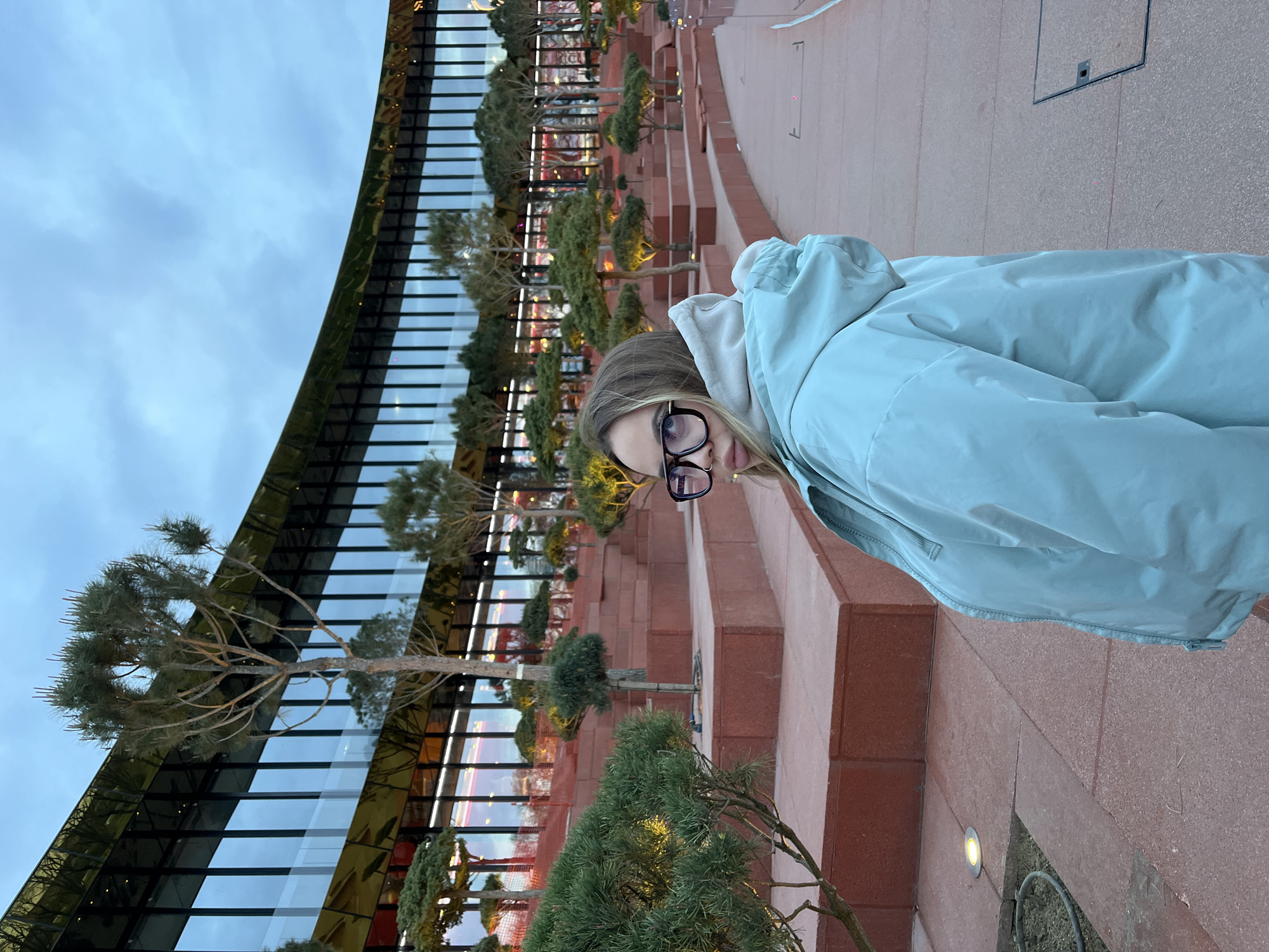 The image captures a young woman posing outdoors in a modern urban landscape in Krasnodar, Russia. The woman, positioned on the right side of the frame, has long, light brown hair and wears black-rimmed glasses. She is dressed in a light blue or mint green hooded jacket layered over a lighter, possibly beige, hoodie, and looks directly at the viewer with a neutral expression.
In the background, a large, contemporary building with a dark, reflective glass facade stretches across the left side of the scene. The facade exhibits an angled or curved design, reflecting a pale, overcast sky. Along the base of this building, a series of reddish-brown, tiered terraces are landscaped with several small, sculpted evergreen trees and bushes, arranged in an orderly fashion. The foreground and pathway areas are also made of the same reddish-brown material, suggesting a cohesive design.
Small, circular recessed lights are visible in the terraced areas and on the ground, indicating they are illuminated. The cloudy, light grey-blue sky suggests either a heavily overcast day or the twilight hours of late afternoon or early evening, which is further supported by the illuminated ground lights. No specific activity beyond posing for the photograph is evident, and there is no visible text in the image. The setting appears to be a well-maintained modern public space or architectural complex.