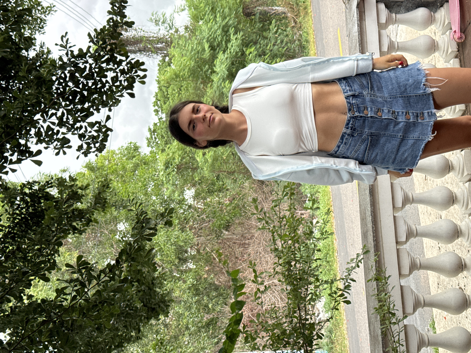 A young woman with dark hair styled in braids stands outdoors, facing the camera with a slight smile. She is wearing a white crop top, a light blue zip-up jacket, and a denim skirt. The background consists of lush green trees and foliage, with a road visible in the distance. The scene appears to be on a porch or balcony with a white railing. There is a pink scooter on the ground to the right of the woman. This location is not identifiable as Mũi Né, Vietnam.