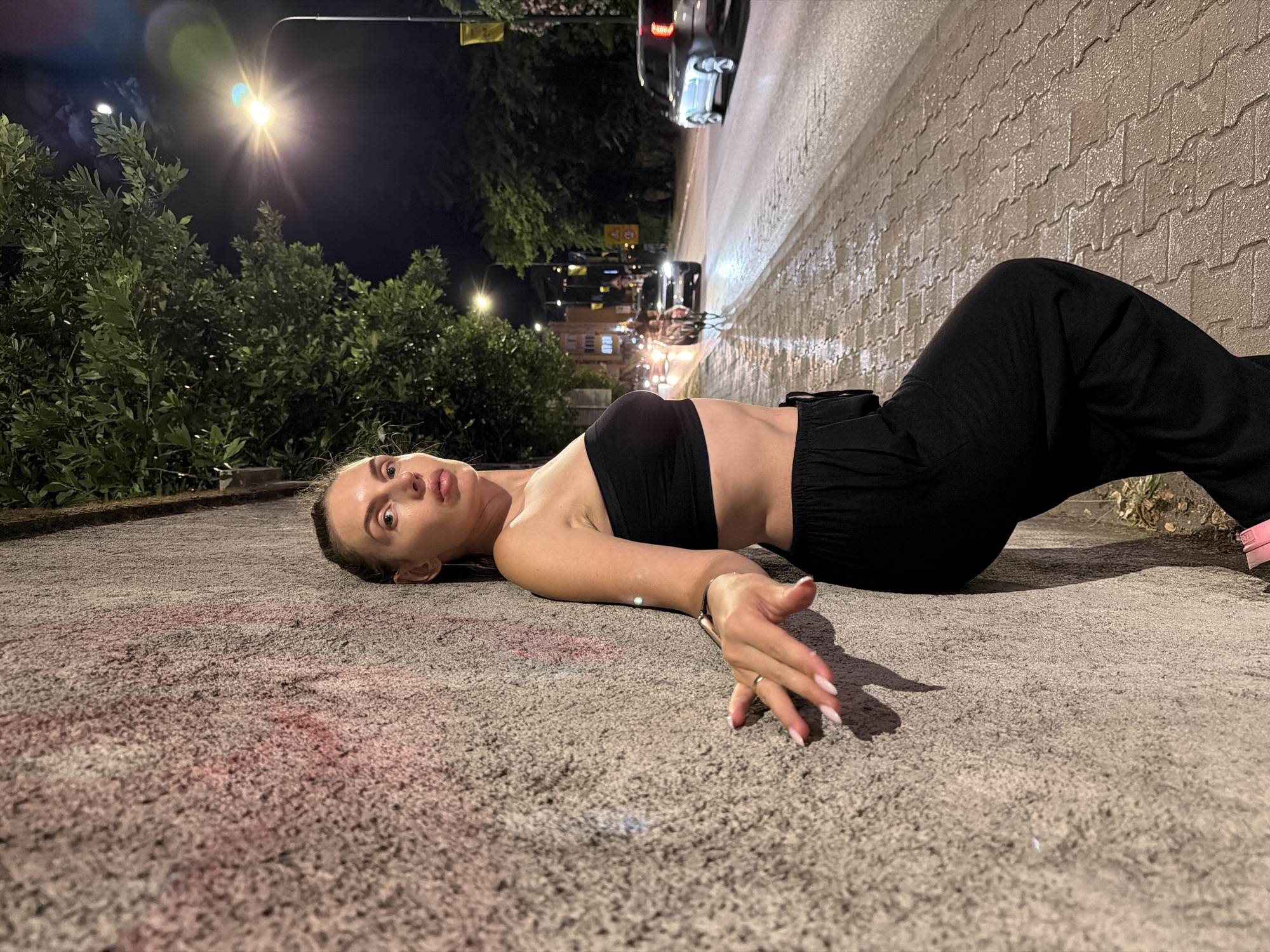 A young woman is lying on her back on a textured concrete surface at night. She is wearing a black crop top and black loose-fitting pants. Her head is turned towards the camera, and she has a surprised or questioning expression on her face with her eyes wide open. Her right arm is extended, with her hand resting on the ground. She is wearing a ring on her ring finger.
The scene appears to be a street or sidewalk at night, with a streetlamp illuminating the area. To the left of the woman are dark green bushes, and further back, there are more trees and some buildings with lights. A street with cars driving on it is visible to her right, and a brick wall runs alongside the street. The concrete surface she is lying on has some reddish-pink marks on it. The time of day is clearly night. The presence of the street, cars, and buildings suggests an urban or semi-urban environment. The lighting from the streetlamp creates distinct shadows.