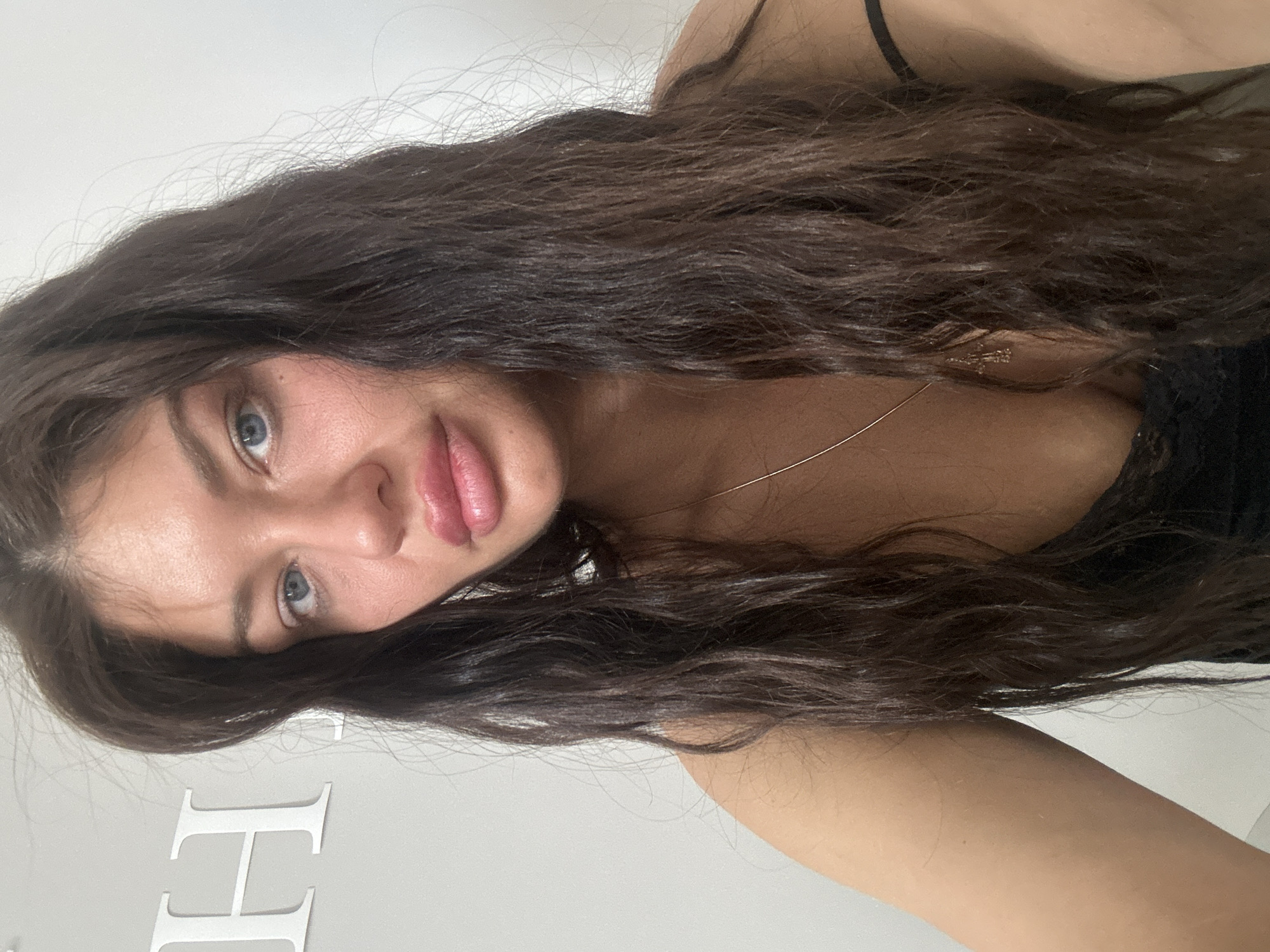 A woman with long, dark brown, wavy hair, blue eyes, and full lips, faces the camera. She appears to be wearing a black top and a delicate necklace. Part of a white wall is visible in the background with large, white, block letters, likely spelling a word or name, extending along the lower left portion of the image. The scene's location in Moscow, Russia is unsubstantiated by the visual evidence.