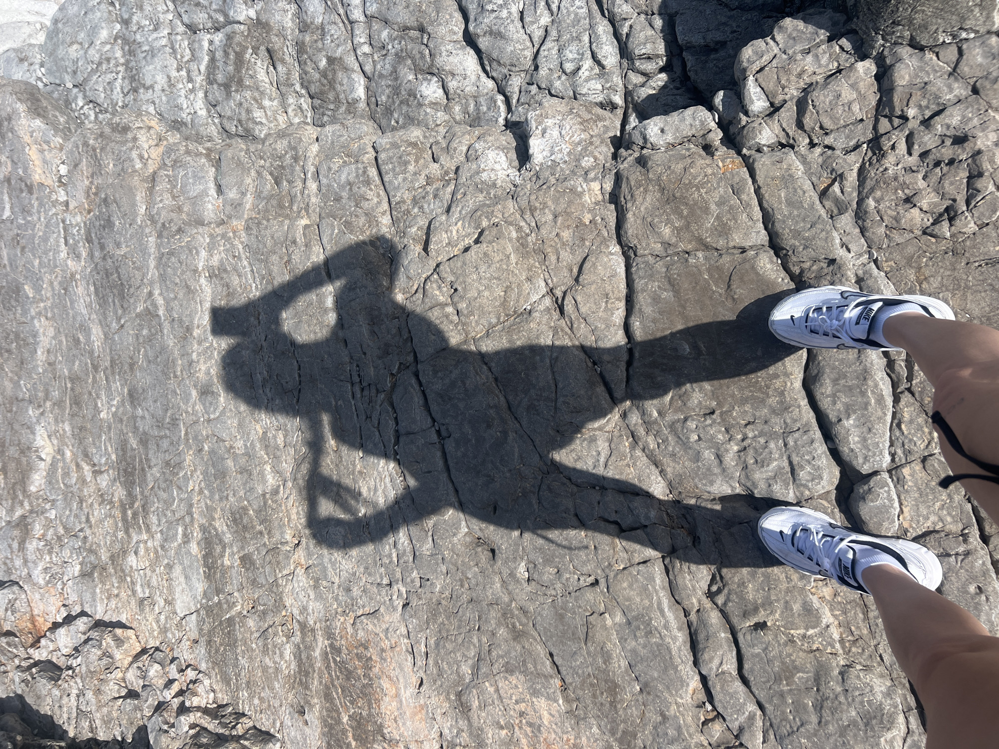 View from above of light grey-colored textured rock surface, possibly weathered limestone, in an outdoor setting. The shadow of a person holding a camera/phone is visible in the center-left of the frame. The legs and feet of the person creating the shadow are in the top and bottom-right of the frame, wearing white running shoes with black accents. The location is Prijevor, Montenegro.