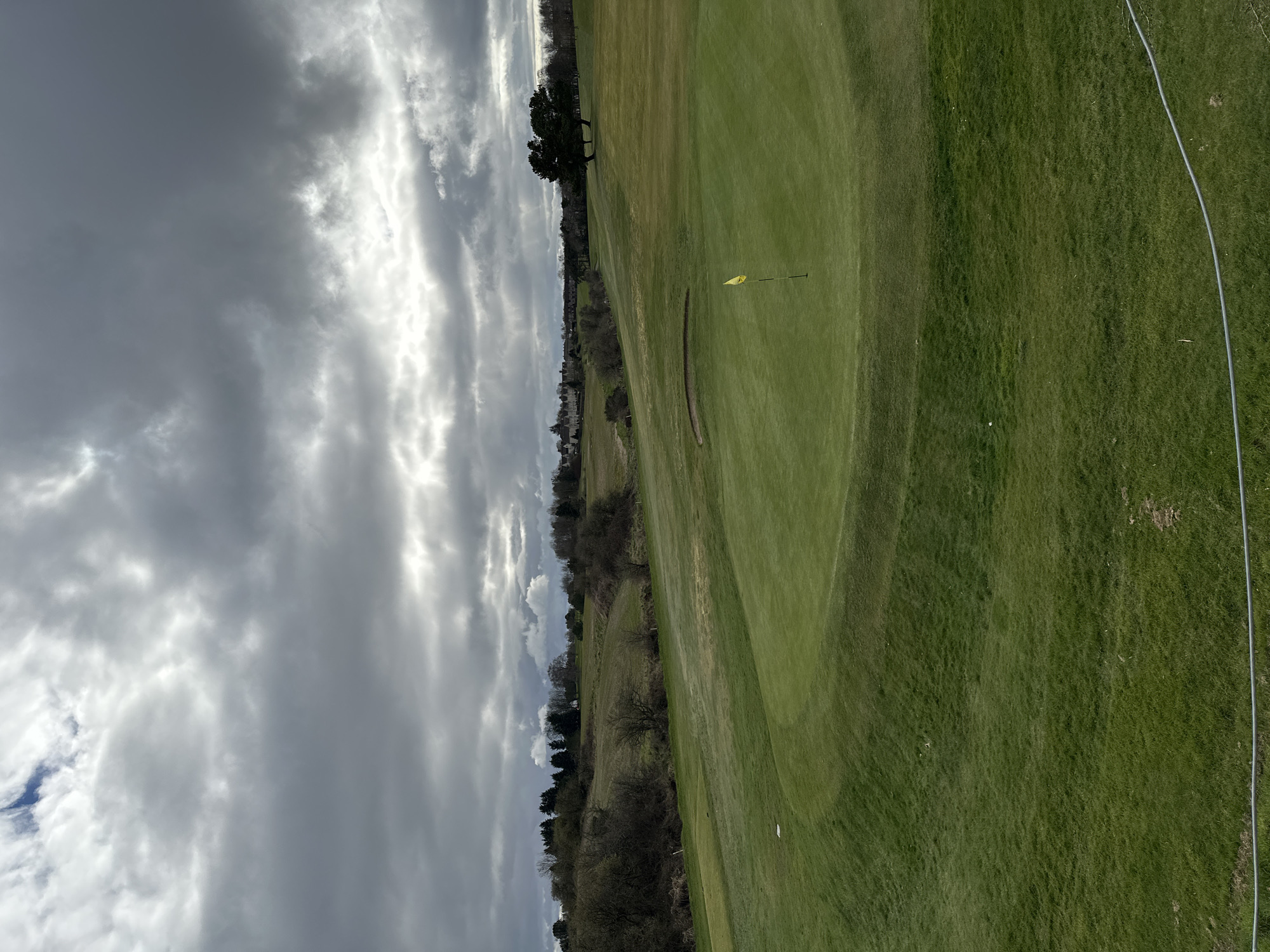 The image depicts a golf course, likely the Pitreavie Golf Club, located in Dunfermline, United Kingdom. The foreground consists of well-maintained green grass, likely part of a fairway or rough. A yellow flag marks the location of a hole on the green. A sand bunker is situated near the green. Beyond the green, there's a line of trees and bushes separating the golf course from an area with some houses or buildings in the distance. The sky is overcast with heavy, textured clouds. A silver fence is running the right side of the image.