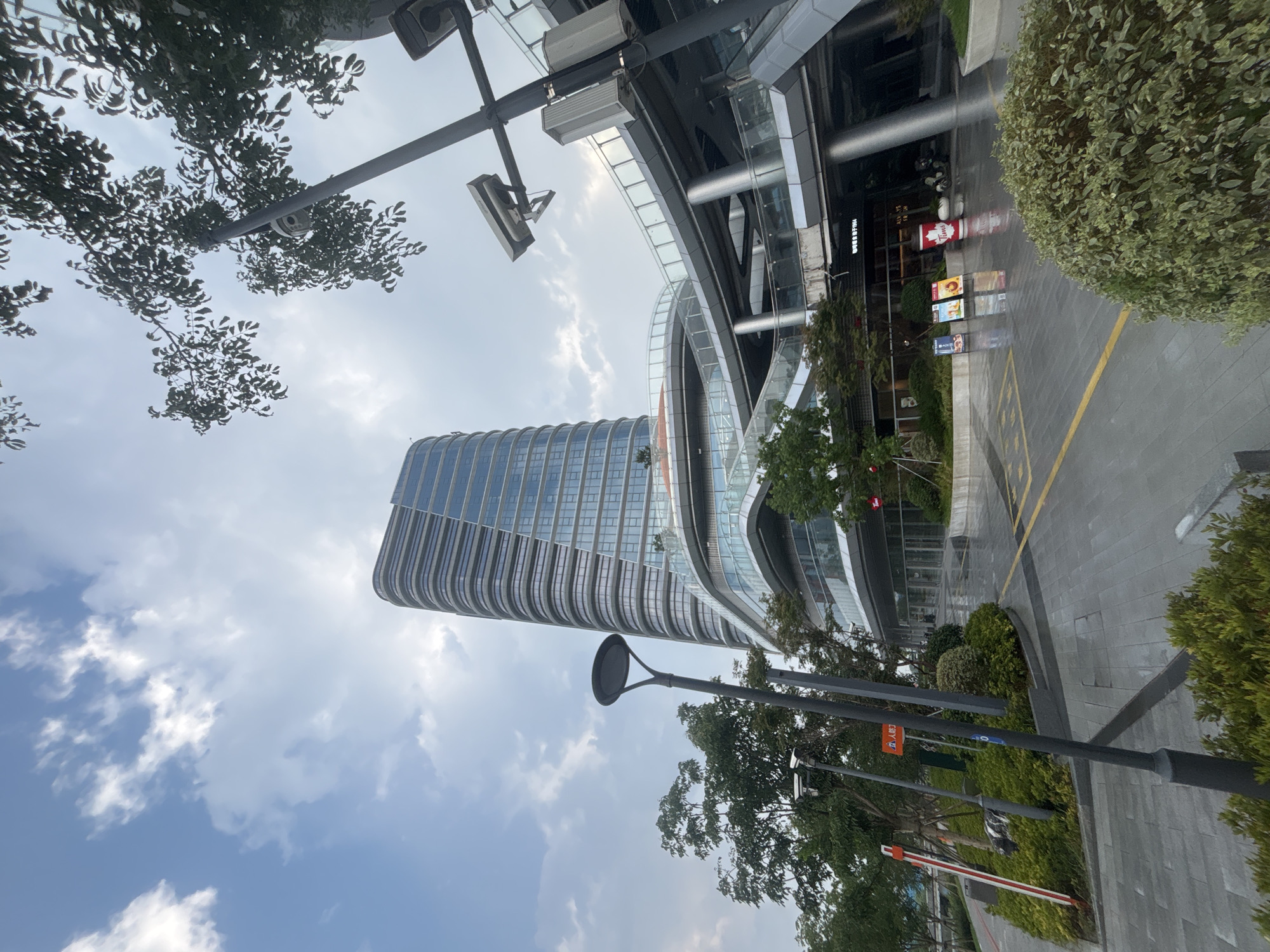 This image captures the exterior of a modern commercial building, likely part of a shopping center or office complex, located in the Suzhou Industrial Park, China. The building features a distinctive, curved architectural design with a tall, multi-story tower that is clad in horizontal bands of glass and metallic material, creating a ribbed effect. Below the tower, several levels of interconnected walkways and balconies, with glass railings and metallic supports, are visible. Lush green trees and manicured bushes surround the building, and the ground in front is paved with light-colored rectangular stones, wet from what appears to be recent rain, with yellow directional lines. Several streetlights, one with a distinctive circular design, are present, along with what appear to be security cameras mounted on poles. In the foreground, to the right, a large, rounded green bush is prominent. In the middle ground, several portable display stands with advertisements and a red cup with the "Tim Hortons" logo suggest food or beverage establishments. A red and white striped barrier, commonly used to control vehicle access, is seen on the left side of the image. The sky above is a mix of blue with scattered white clouds. There are no people actively engaged in the scene.