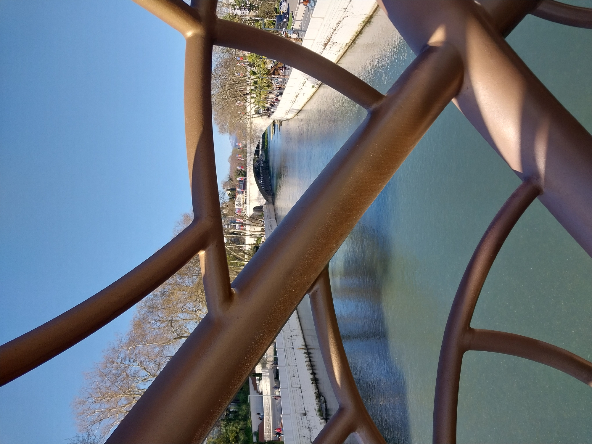The image, taken in Sochi, Russia, presents an outdoor urban scene viewed through an ornate, bronze-colored metal railing or balustrade. This intricate railing, characterized by its interconnected, rounded bars, dominates the foreground, with strong sunlight casting prominent shadows across its surfaces.
Beyond the railing, a calm river or canal flows, its tranquil surface reflecting the bright, clear blue sky overhead. Both banks of the river feature developed infrastructure. The left bank reveals a promenade where several people are seen strolling. The vegetation consists of a mix of lush green palm trees and deciduous trees with bare branches, indicative of a seasonal landscape. Further along this bank, buildings and a road with parked cars are visible, alongside flags displayed on poles in the distance.
Downstream, a bridge gracefully spans the river, connecting the two sides. The right bank also features an embankment or walkway, with additional people enjoying the pleasant weather. The sky is uniformly bright and cloudless, and the distinct shadows suggest either morning or late afternoon on a sunny day. The overall impression is one of a serene and well-maintained public space in a resort city, with no discernible text present in the image.