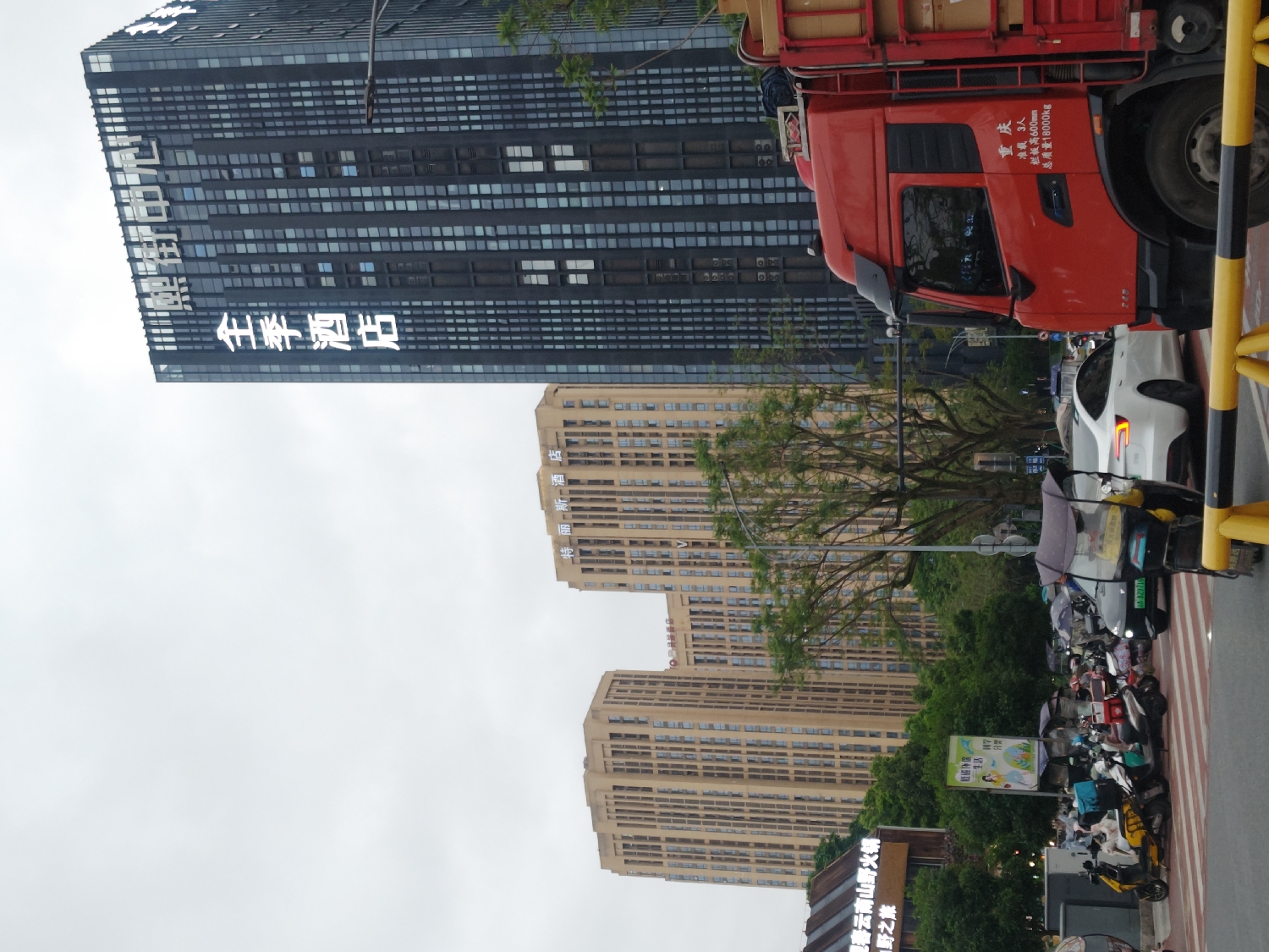 The image depicts a bustling urban street scene in Chongqing High-tech Zone, China, under an overcast sky during the daytime. The perspective captures a mix of modern high-rise buildings, various vehicles, and people going about their daily activities.
A prominent dark, modern high-rise building with a glass facade stands on the left side of the frame, featuring large, illuminated white Chinese characters spelling "全季酒店" (Quanji Hotel). In the middle and background, several light brown residential or commercial high-rise buildings are visible, one of which displays "特丽斯酒店" (Telisi Hotel) on its upper section.
The foreground showcases street activity. A large red truck with a cargo bed is prominently positioned on the right. Text on its side indicates "重庆" (Chongqing), "准载 3人" (Approved load 3 people), "栏板高600mm" (Side panel height 600mm), and "总质量18000kg" (Gross mass 18000kg). A white car, possibly a Tesla, is parked behind the truck. Along the roadside, numerous motorcycles and electric scooters are parked, many equipped with umbrellas for sun or rain protection. A partial license plate "ABZ9716" is visible on one scooter. Several people are seen near the parked vehicles, some holding umbrellas. A yellow safety barrier separates the street from the pedestrian and parking area.
Amidst trees and other structures further down, a sign for "夏春云南山野火锅" (Xiachun Yunnan Mountain Wild Hot Pot) is visible, along with "野之旅" (Wild Journey/Tour) and "生活" (Life). The scene portrays a typical busy day in a modern Chinese city, characterized by a blend of commercial establishments, residential areas, and active street life.