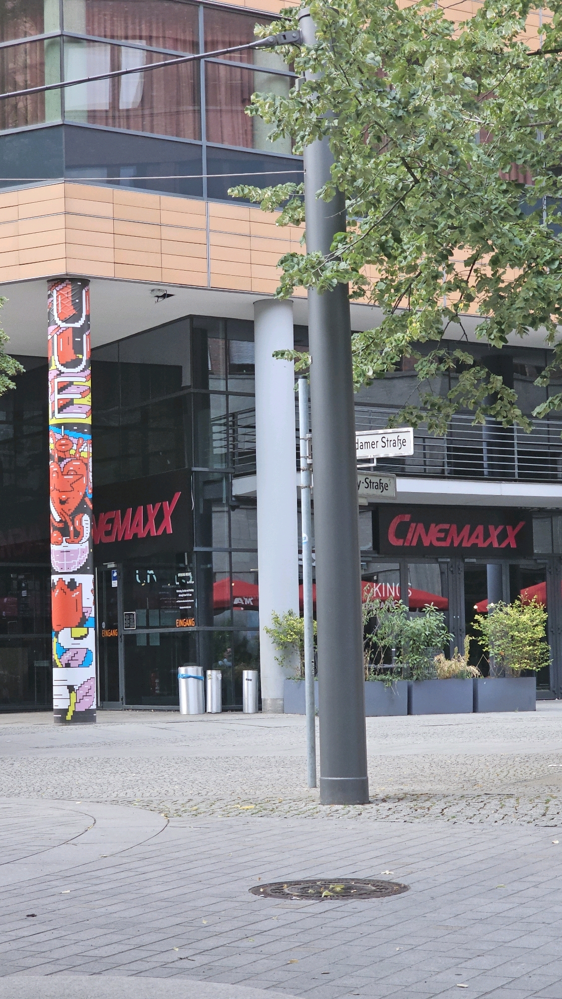 The image shows the exterior of a Cinemaxx cinema located at Dammer Straße in Berlin, Germany. A modern building with large glass windows and an orange facade forms the backdrop. In the foreground, a tall, grey lamppost stands prominently. To the left, a cylindrical pillar is decorated with colorful pixelated artwork featuring a red heart. Two entrances labeled "EINGANG" are visible. Red umbrellas are set up outside the cinema. In front of the building, there are several modern cylindrical trash cans. The ground is paved with grey interlocking stones, and a manhole cover is visible in the lower right quadrant. A tree with green foliage is partially obscuring the building on the right side of the frame.