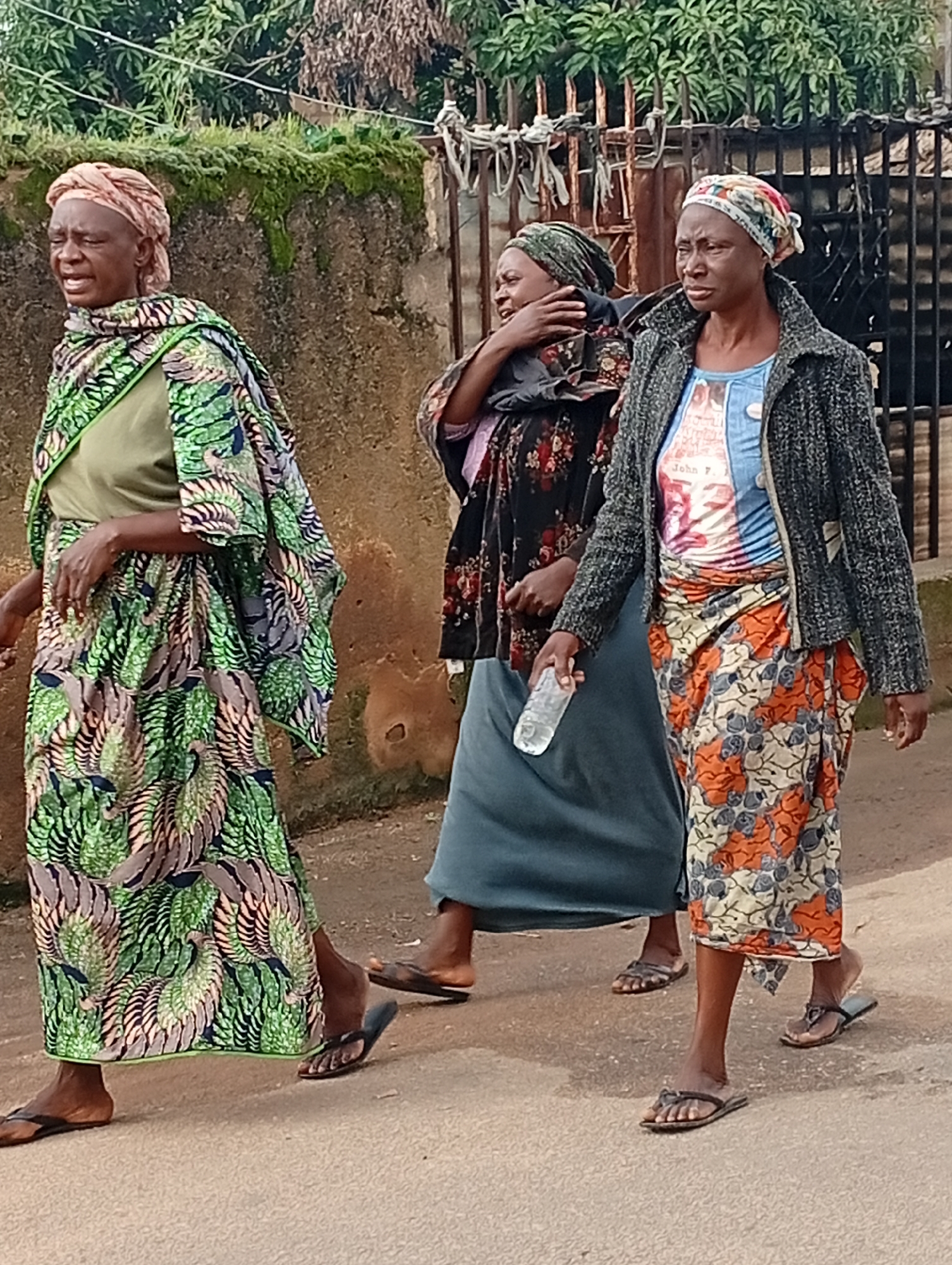 In Jos, Nigeria, three women are walking outdoors on a dusty path with a rough-textured, moss-covered wall behind them and a black metal fence to the right. The woman on the left is wearing a light pink headscarf, a light green garment, and a long, colorful green and black patterned wrap. She is looking to her left with her mouth open, as if speaking or expressing emotion. The middle woman is wearing a dark headscarf and a black and red floral patterned dress, with her left arm raised and hand near her face. The woman on the right is wearing a colorful patterned headscarf, a grey knitted jacket over a colorful t-shirt, and a long orange, white, and blue patterned wrap skirt. She is looking forward, holding a clear plastic water bottle in her right hand. All three women are wearing open-toed sandals. The ground is a light brown, textured surface, suggesting a dirt or unpaved road. The environment appears to be a residential or semi-urban area.