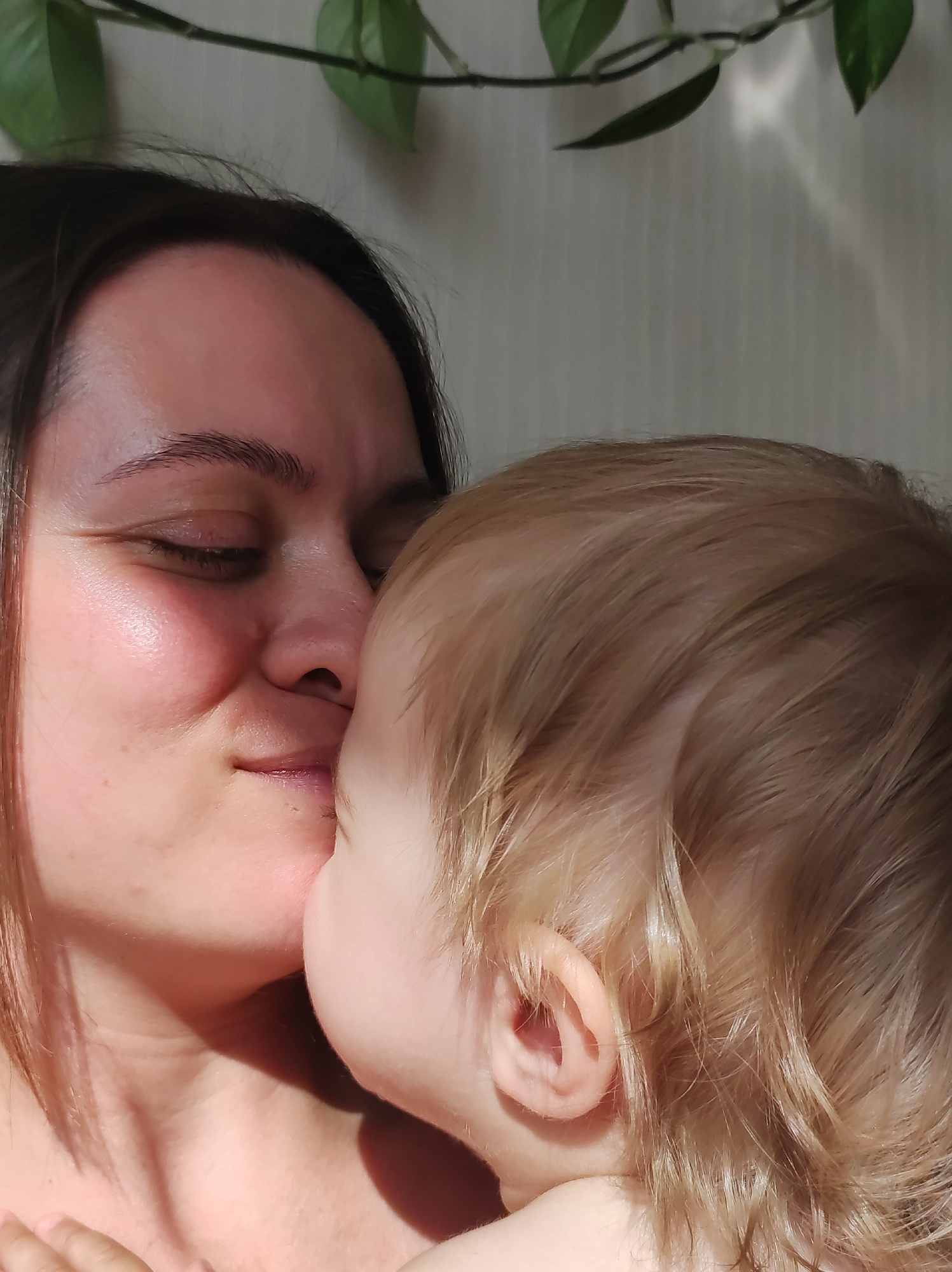 The image features a close-up of a woman holding a young child. The woman has dark hair and her eyes are closed. The child has blond hair. Their faces are close, possibly touching. A green plant is visible in the background, situated behind them. The background appears to be a plain white wall with some light or shadow patterns. There is no evidence to suggest the location is Chaikovsky, Russia.
