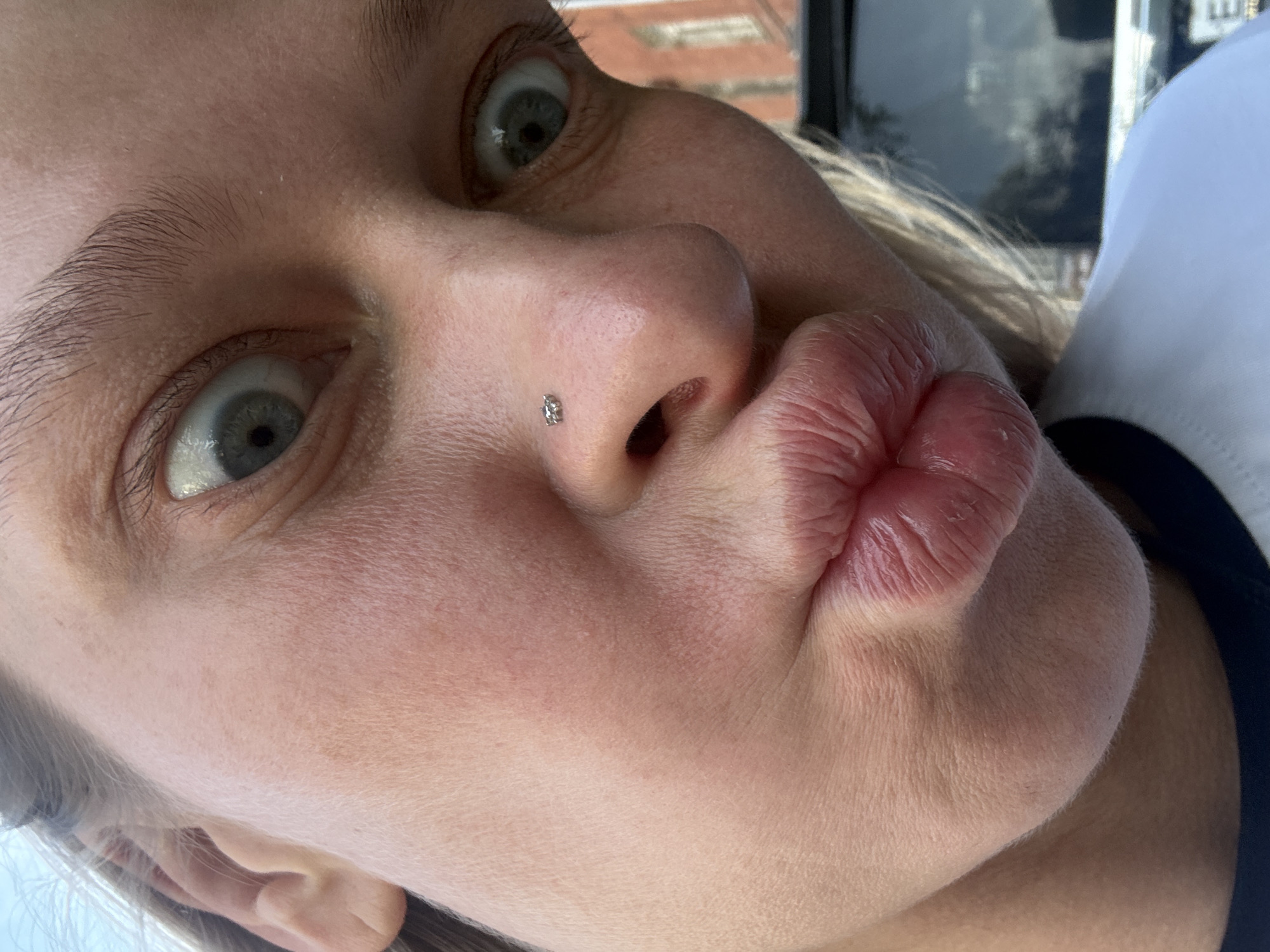 A close-up, somewhat distorted, selfie of a light-skinned person with blonde hair is presented. The face is angled towards the camera, with wide, expressive eyes and pursed lips in a "kissing" expression. A small stud is visible on the left nostril. The background is blurred, showing indistinct shapes that might be buildings and foliage. It is not possible to determine the location as Perm, Russia, based on visual data provided.