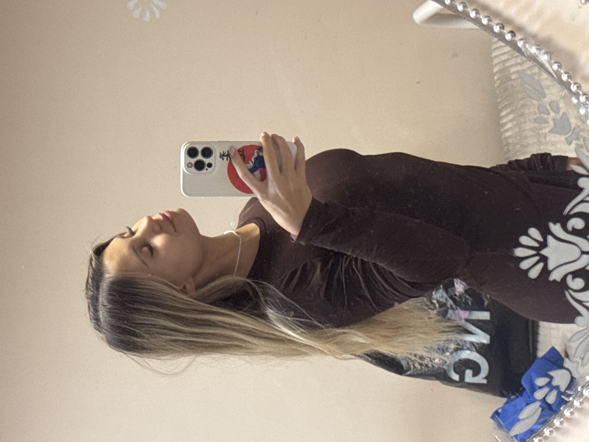A woman with blonde hair is taking a selfie in a mirror. She's holding a smartphone with a white case and a graphic on the back. She wears a long-sleeved brown garment. The mirror has a decorative beaded frame. The background includes a beige wall and decorative elements. The location, Chaikovsky, Russia is not visually determinable from this image.
