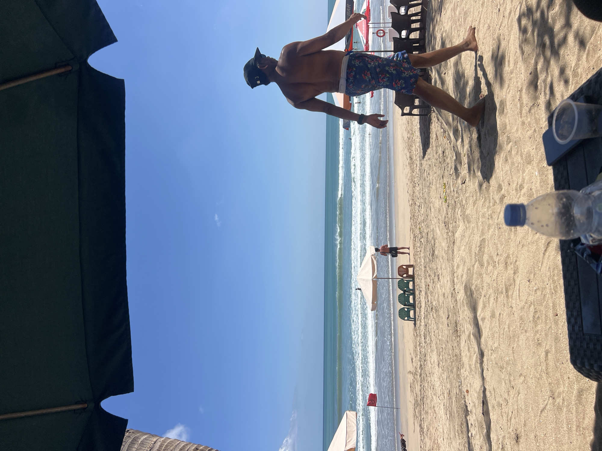 The image depicts a sunny day at Kuta Beach in Kuta, Indonesia. In the foreground, a table with a partially visible plastic cup, a smartphone, and a water bottle is in the shade of an umbrella. The sand is textured with shadows cast by unseen objects. In the midground, a man wearing a black baseball cap and floral swim trunks walks from right to left, his back to the camera. He is shirtless and appears to be in motion, with his right leg mid-stride. Behind him, the beach stretches towards the ocean. Several beach umbrellas and rows of empty chairs are set up on the sand. A red flag on a pole is planted near the water's edge, indicating beach conditions. The ocean is visible with gentle waves rolling towards the shore. In the distance, the horizon meets the clear blue sky. Another person, smaller in the frame, stands near the water further down the beach.