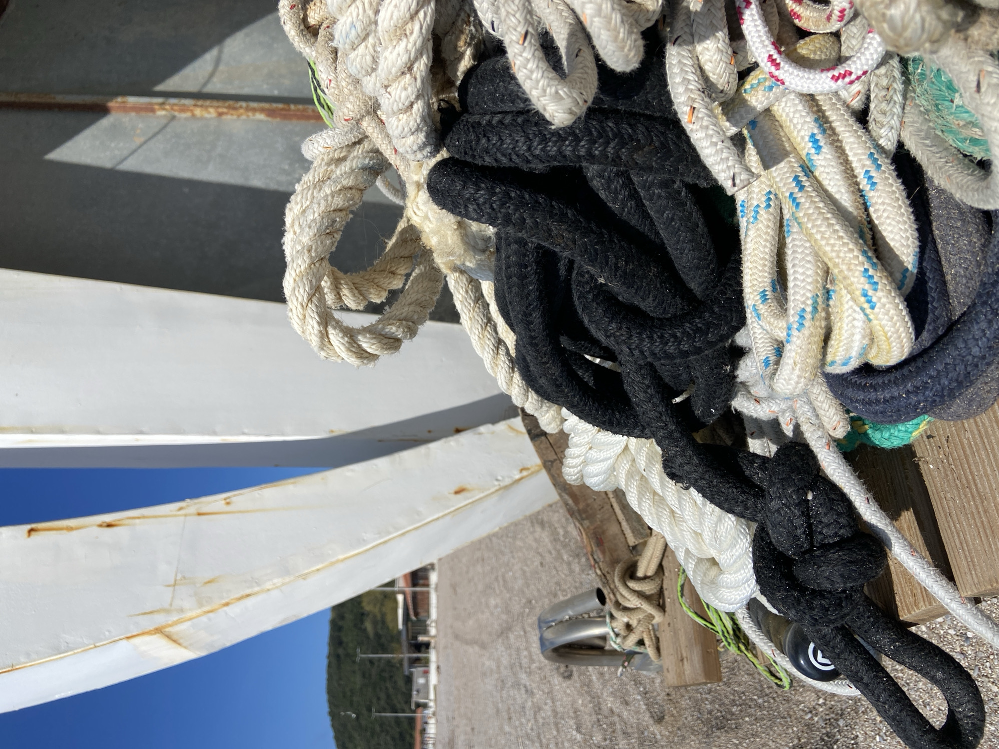 The image shows a close-up of assorted ropes, mostly white and black, some knotted. The ropes are piled on a wooden surface. The edge of a metal structure is visible, along with a glimpse of the town of Prijevor, Montenegro, featuring buildings and trees under a blue sky. The location appears to be near the waterfront.