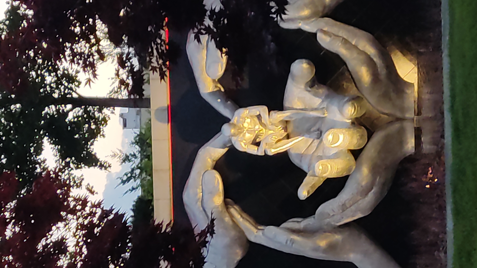 This is a vertical, slightly blurry, low-light photograph of a sculpture depicting human hands cradling a smaller figure. The sculpture appears to be made of metal and is illuminated, highlighting its contours. The hands are arranged in a circular formation, with one pair above and another pair below, creating a protective embrace around the central figure. The central figure is abstract and appears to be composed of metallic elements as well, possibly representing a person or a symbolic object. The overall impression is one of care and support.
In the background, out of focus, are hints of a natural setting. Dark foliage, possibly from trees with red leaves, frames the upper left portion of the image. Beyond the foliage, there's a glimpse of a building or structure and the sky, suggesting an outdoor location. The lighting and the dark tones indicate it is either dusk or a heavily shaded area. There are no visible text elements in the image. The scene evokes a sense of tenderness and protection.