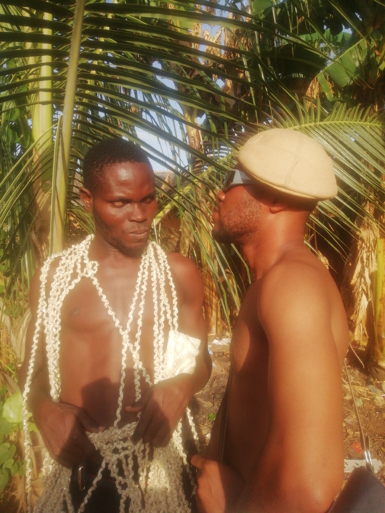 Two dark-skinned men stand outdoors amidst lush tropical vegetation. The man on the left is shirtless and draped in multiple strands of white, rope-like material resembling a garment. He gazes intently towards the man on the right. The man on the right wears a light tan cap and sunglasses, his torso bare. The background includes green palm fronds and other dense foliage. The location is the city of Unknown, Unknown.