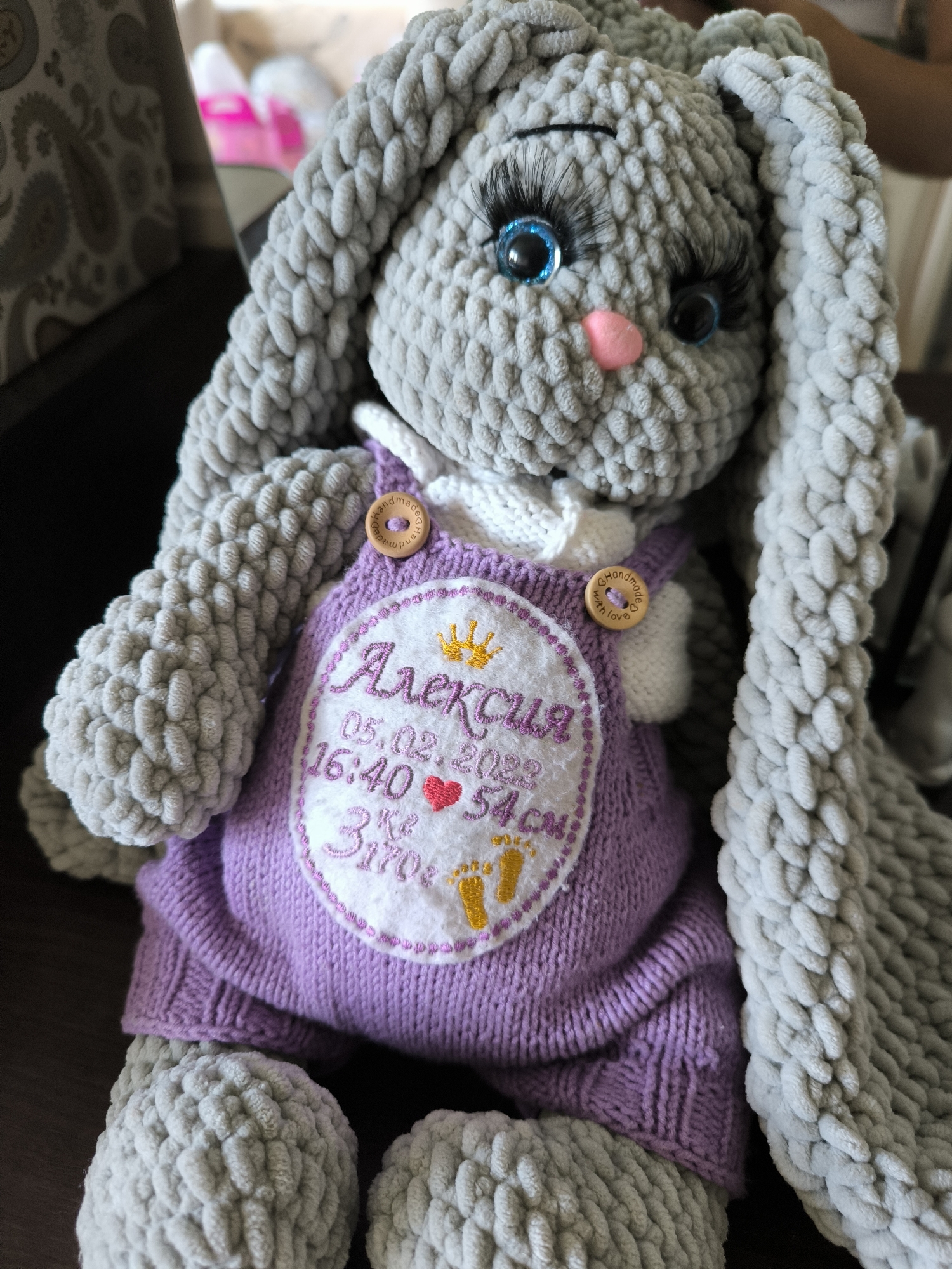 A handmade, grey crocheted bunny toy is the central subject. It is wearing a purple knitted romper with a white embroidered oval patch. The patch contains text and graphics in pink and yellow thread. At the top of the patch is a golden crown, below which reads "Алексия". Beneath the name are the date "05.02.2023", the time "16:40", a red heart, and the text "3 кг 3170 г 54 см". Two small wooden buttons with "Handmade with love" inscribed on them are attached to the romper straps. The bunny has large, bright blue eyes with long black eyelashes and a small pink nose. It is sitting on a dark brown surface, possibly a table, with a patterned wallpaper visible in the blurred background to the left. The image's perspective is a close-up, slightly angled view from the front. The location is not specified in the image.