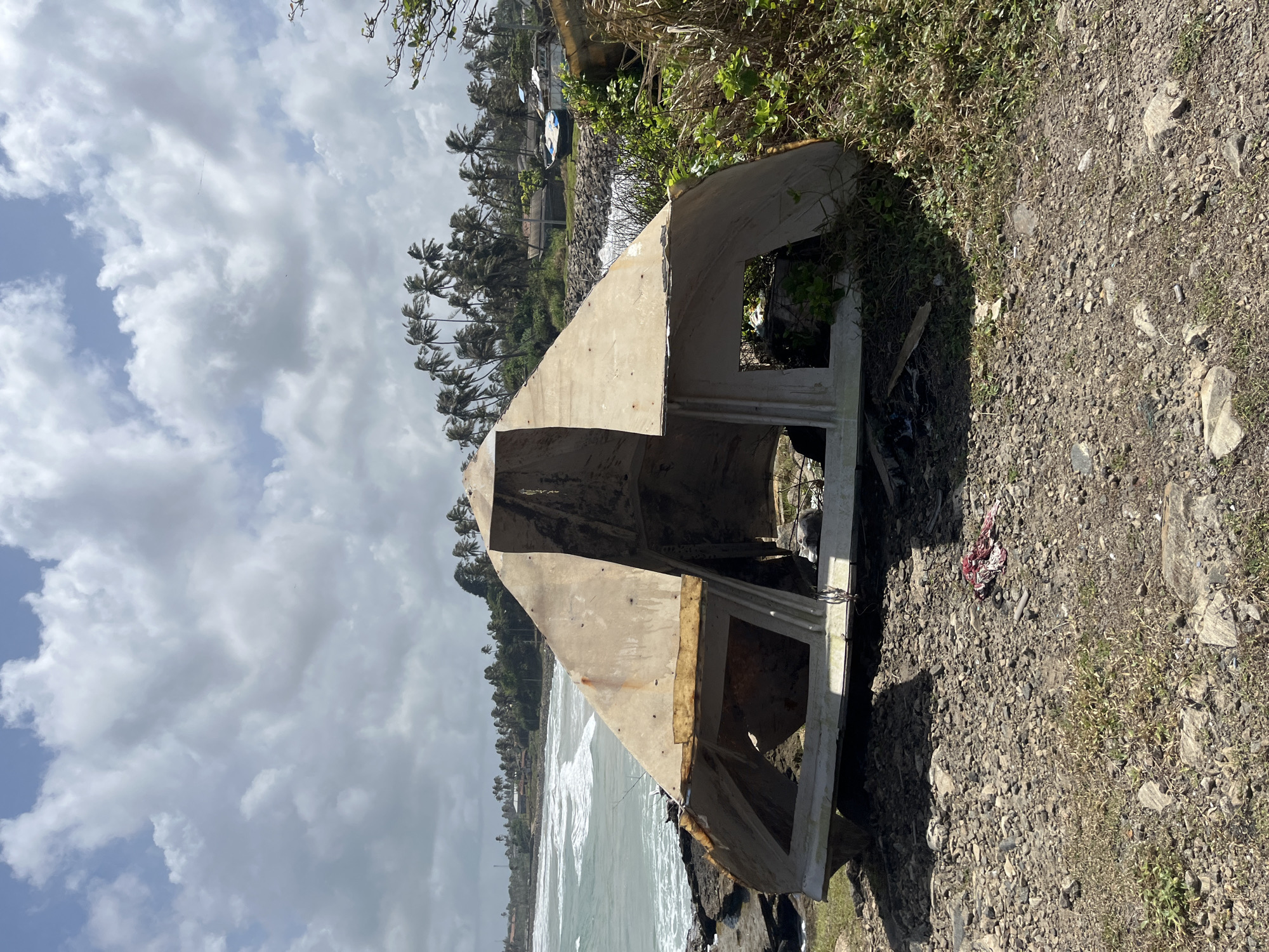 The image depicts a derelict, overturned boat or boat hull on a rocky shoreline, possibly in Dondra, Sri Lanka. The structure is made of weathered, light-colored material, likely fiberglass or wood, and appears damaged and abandoned, with openings revealing an interior filled with shadows and some vegetation. The surrounding environment includes a rough, gravelly ground with sparse grass and scattered rocks in the foreground. In the background, a turbulent sea with white-capped waves meets a rocky coastline. A line of palm trees stretches across the horizon, indicating a tropical or subtropical location. Further inland, several buildings and other boats, some also appearing weathered, are visible. The sky is cloudy, with patches of blue sky visible, suggesting it is daytime. No people are present in the immediate vicinity of the boat.