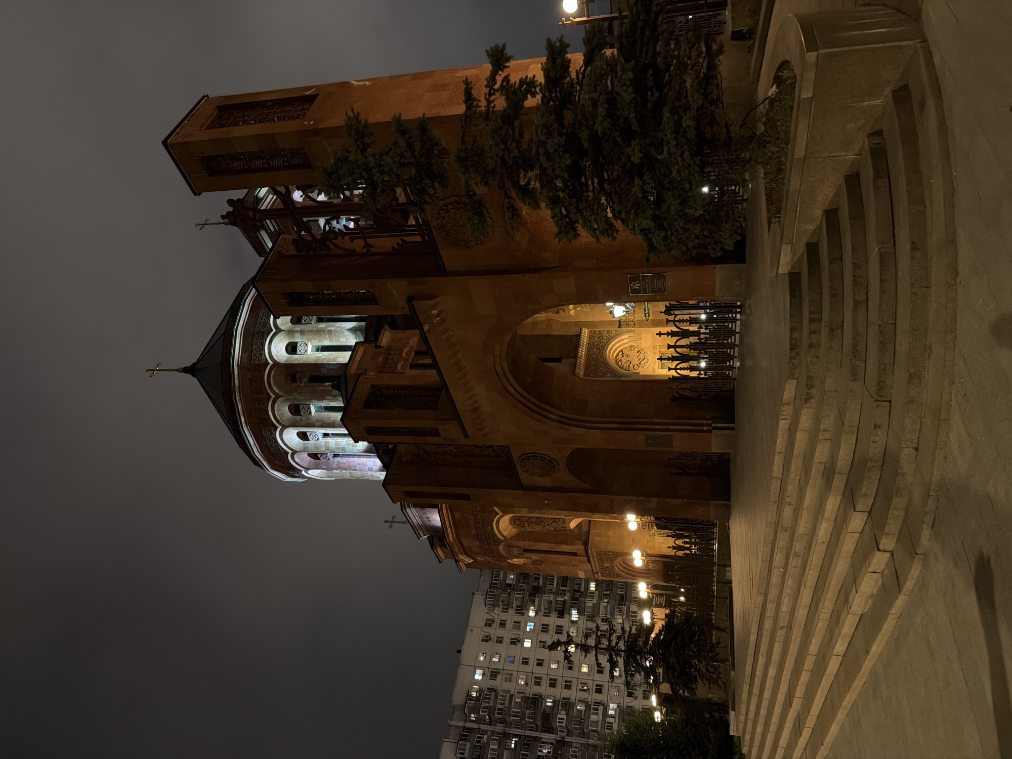 The image shows the Armenian Apostolic Church in Moscow, Russia, at night. The church is illuminated, contrasting with the dark sky. The building is constructed from brown stone and features a tall bell tower, a dome, and arched entrances. A tall apartment building is visible in the background. To the right of the church are stone steps leading up to the entrance. A wrought iron fence runs along the bottom of the stairs. Streetlights provide additional illumination to the area. There is no visible activity or individuals in the shot.