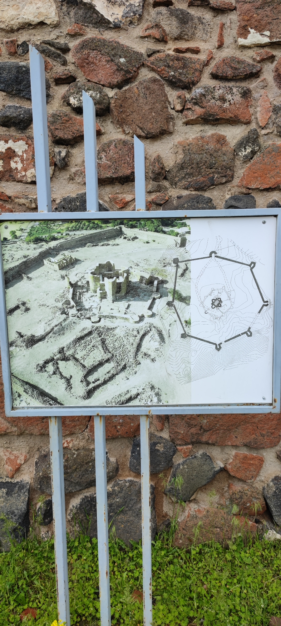 The image shows a signpost in front of a stone wall in Dashtadem, Armenia. The signpost, framed in grey metal, presents information about a historical site, with an aerial view photograph and a schematic map displayed side-by-side. The photograph shows the ruins of a fortress or ancient structure, revealing walls, towers, and other architectural elements. The map illustrates the layout of the same site, outlining the perimeter and key features with contour lines indicating the topography. The wall behind the signpost is constructed from irregularly shaped stones of varying sizes and colors, predominantly shades of brown, grey, and red. In the foreground, green vegetation, including grass and small flowering plants, grows at the base of the wall and around the supporting metal posts of the sign. The scene is outdoors, likely in an area near the historical site represented on the sign.